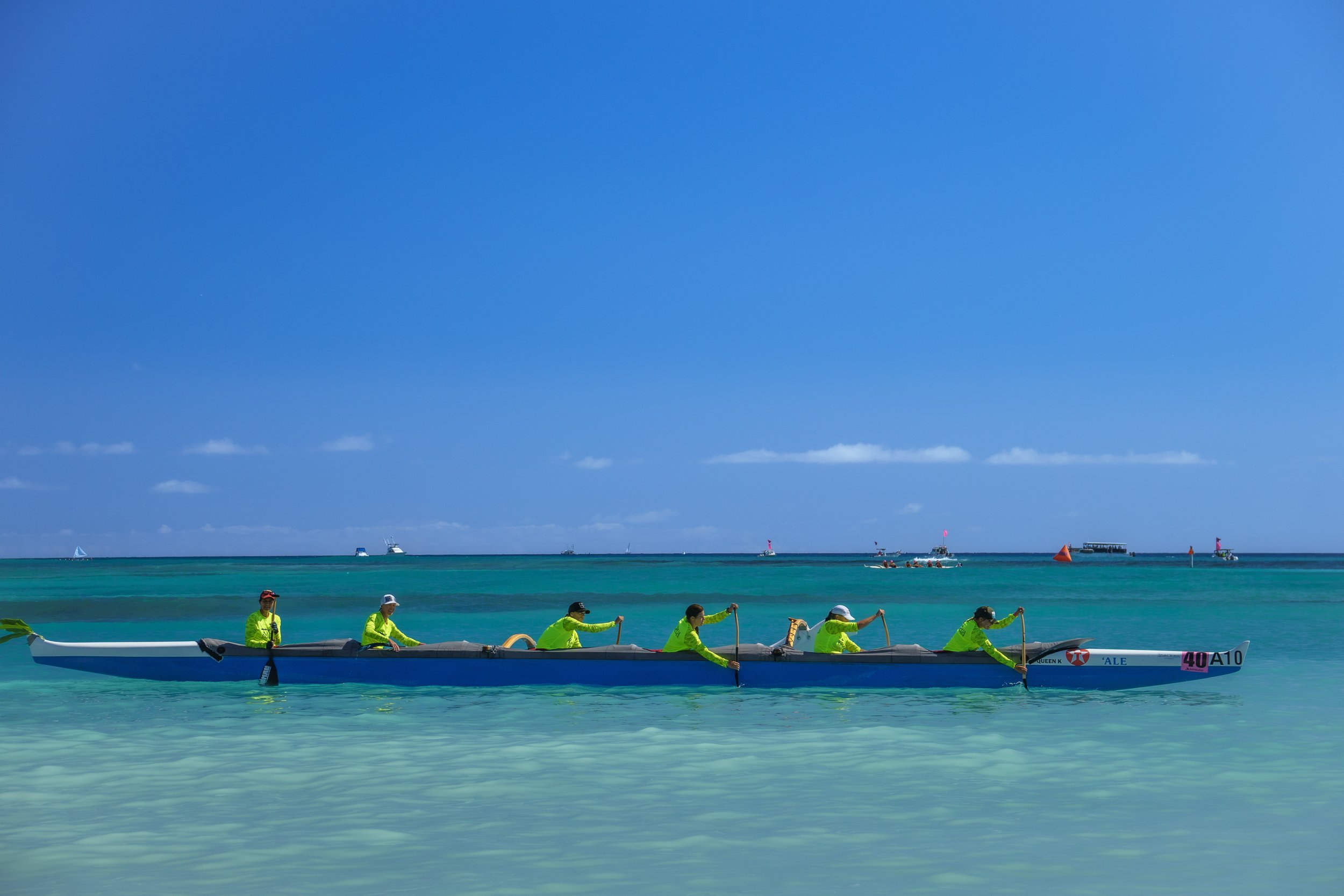 A team of people wearing yellow shirts paddling an outrigger canoe in clear blue water under a bright blue sky.