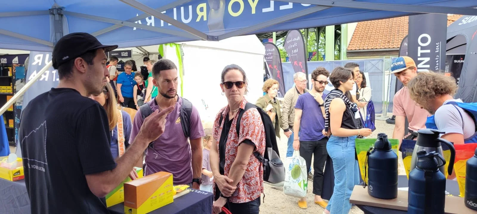 Groupe de personnes à un stand d'exposition lors d'un événement en plein air, avec des affiches et des brochures, certains discutant, d'autres regardant les produits.