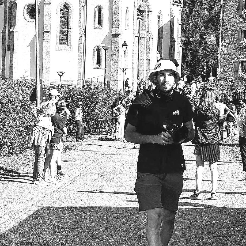 Une photo en noir et blanc d'un jeune homme portant un casque et tenant un appareil photo, dans une foule de personnes devant une vieille église ou un bâtiment historique, avec des buissons et des lampadaires le long du chemin.