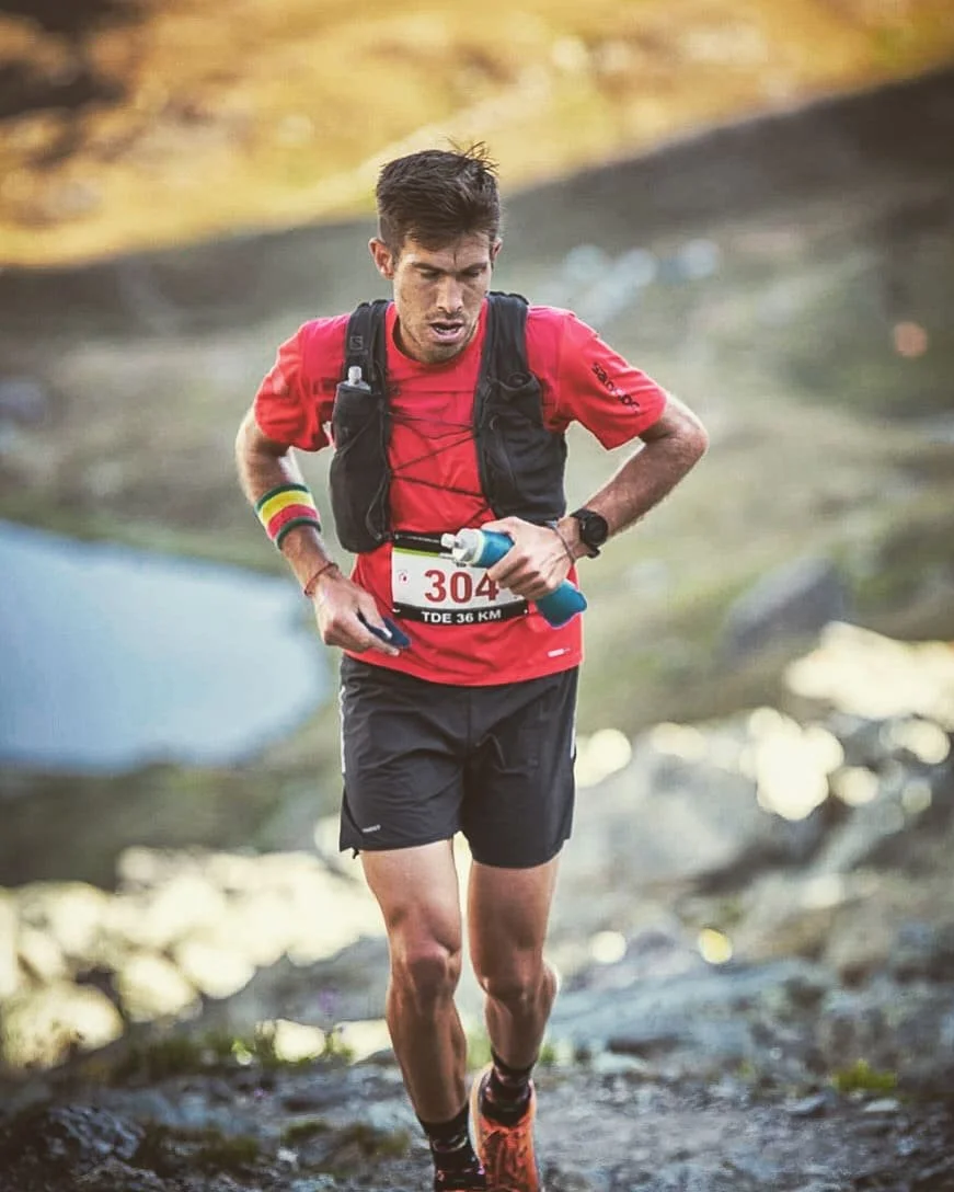 Un homme courant en pleine nature portant une tenue de trail et un sac à dos, avec un bracelet coloré au poignet gauche.