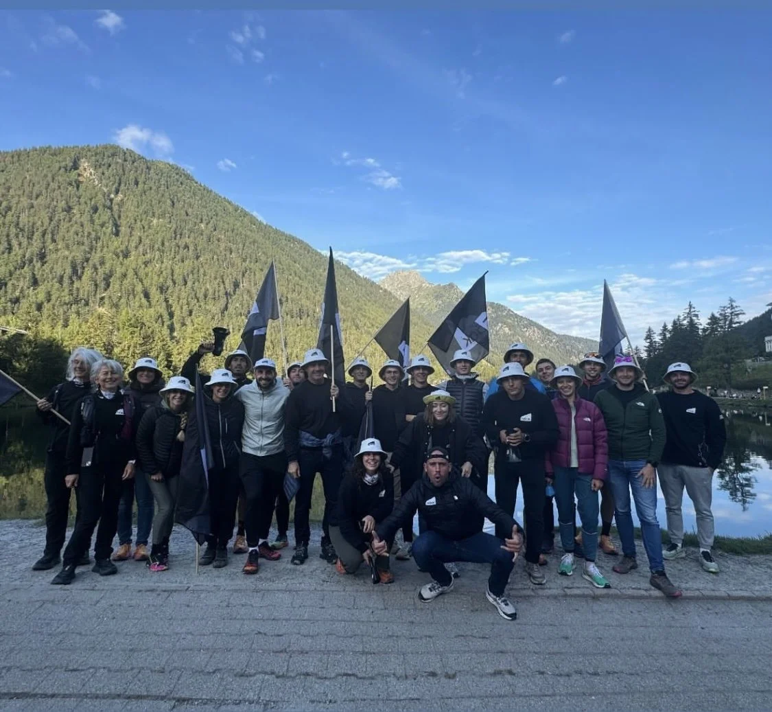 Groupe de personnes posant avec des drapeaux dans un cadre montagnard, portant des chapeaux blancs, par une journée ensoleillée.