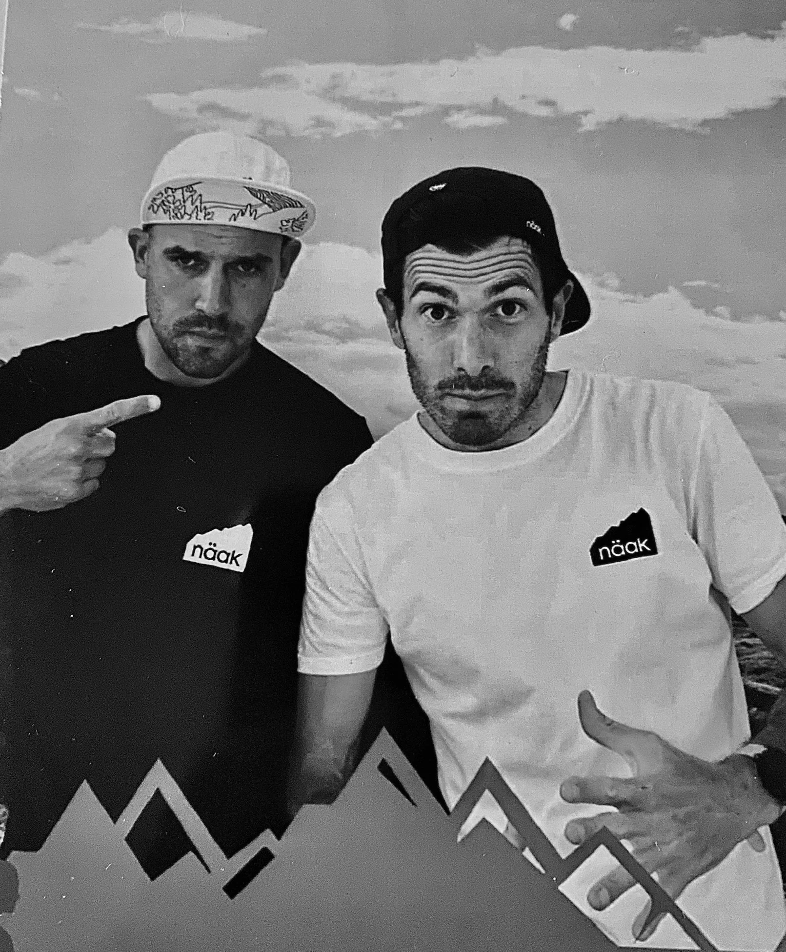 Deux hommes posant avec des expressions sérieuses, l'un pointant son doigt vers l'autre, portant tous deux des t-shirts avec le mot 'näak'.