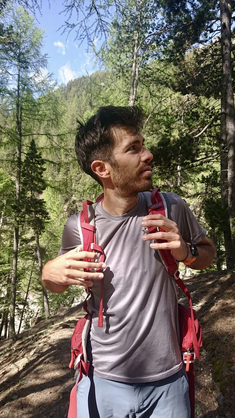 Un homme avec un sac à dos dans une forêt, regardant vers la droite, en pleine nature avec des arbres verts et un ciel bleu.
