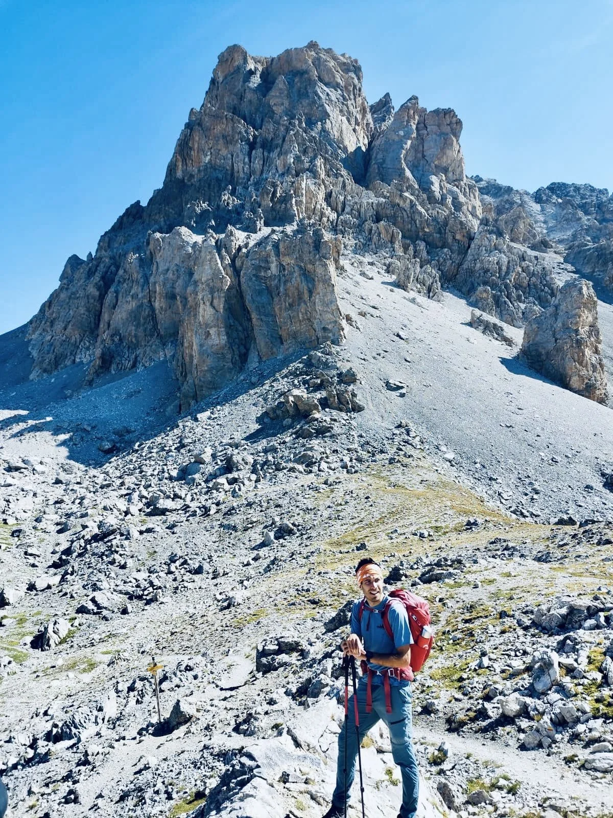Un homme avec un sac à dos et une canne de marche en montagne rocheuse avec un grand sommet en pierre sous un ciel bleu.
