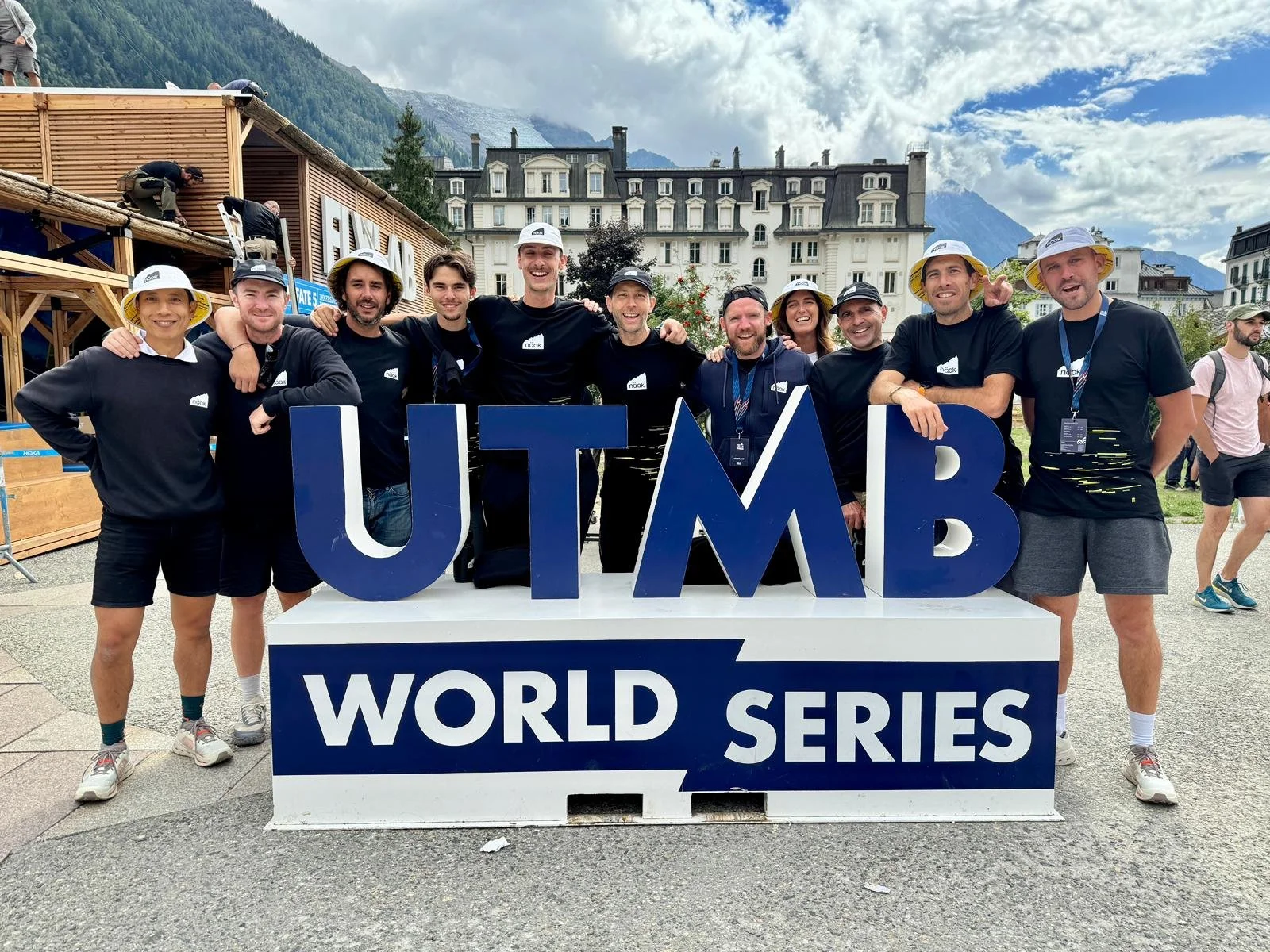 Groupe de personnes posant derrière une grande enseigne 'UTMB WORLD SERIES' dans un cadre montagneux, groupe souriant portant des t-shirts noirs, certains portant des chapeaux blancs