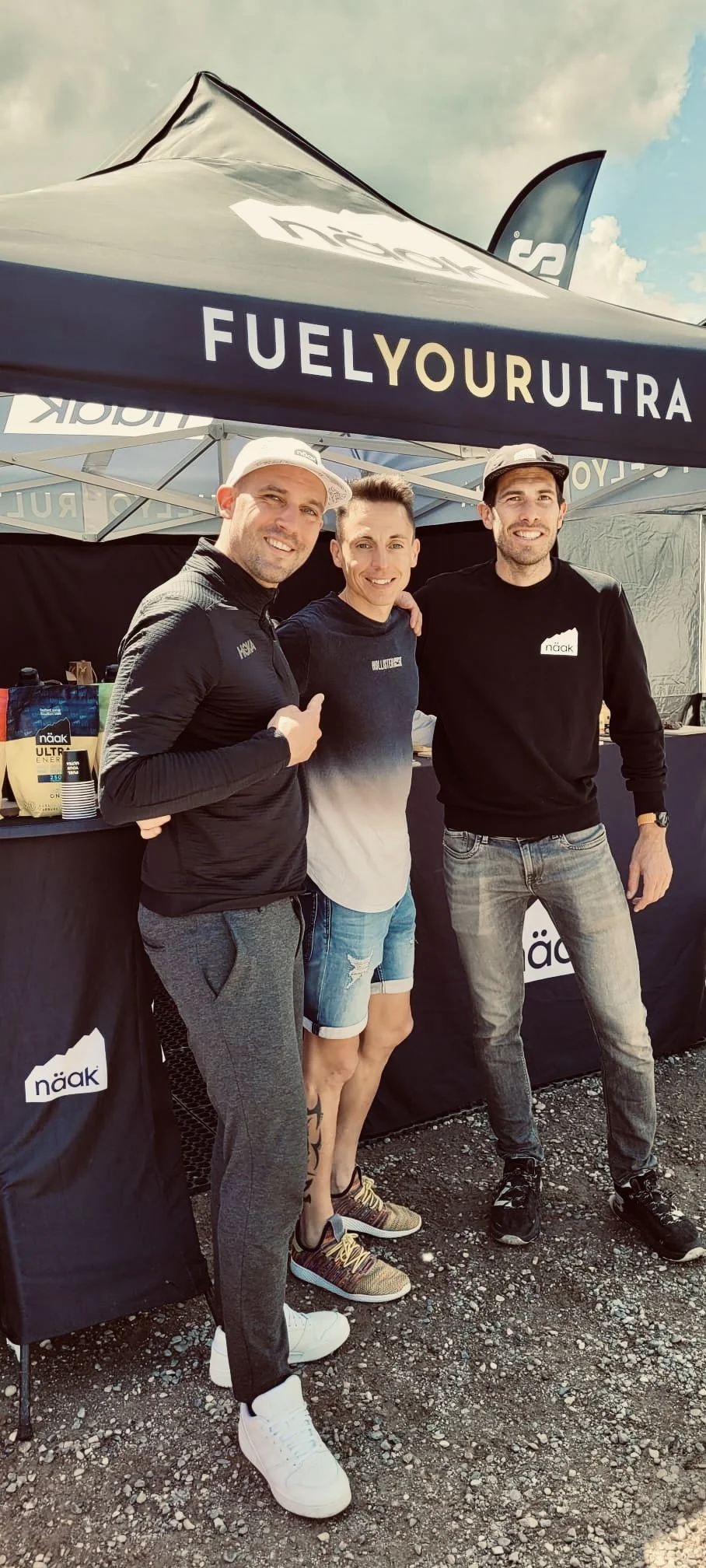 Trois hommes posent ensemble devant un stand avec une tente noire portant le slogan 'FUEL YOUR ULTRA'. Ils sourient et portent des vêtements de sport. Les vêtements du premier homme indiquent 'HOKA', ceux du troisième 'näak'.