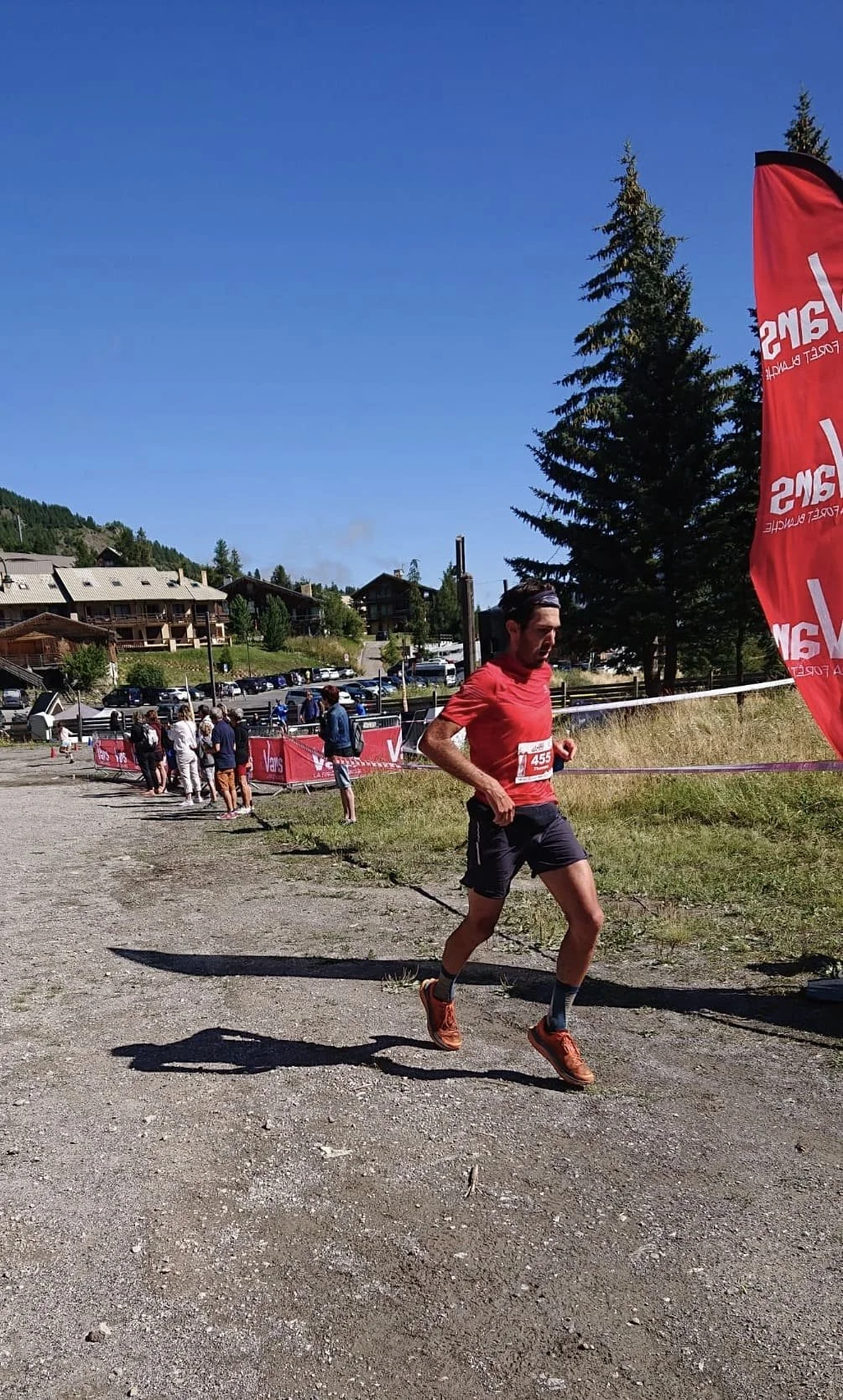 Un coureur participe à une course en plein air sous un ciel dégagé, avec des spectateurs en arrière-plan, en montagne, près de bâtiments en bois.