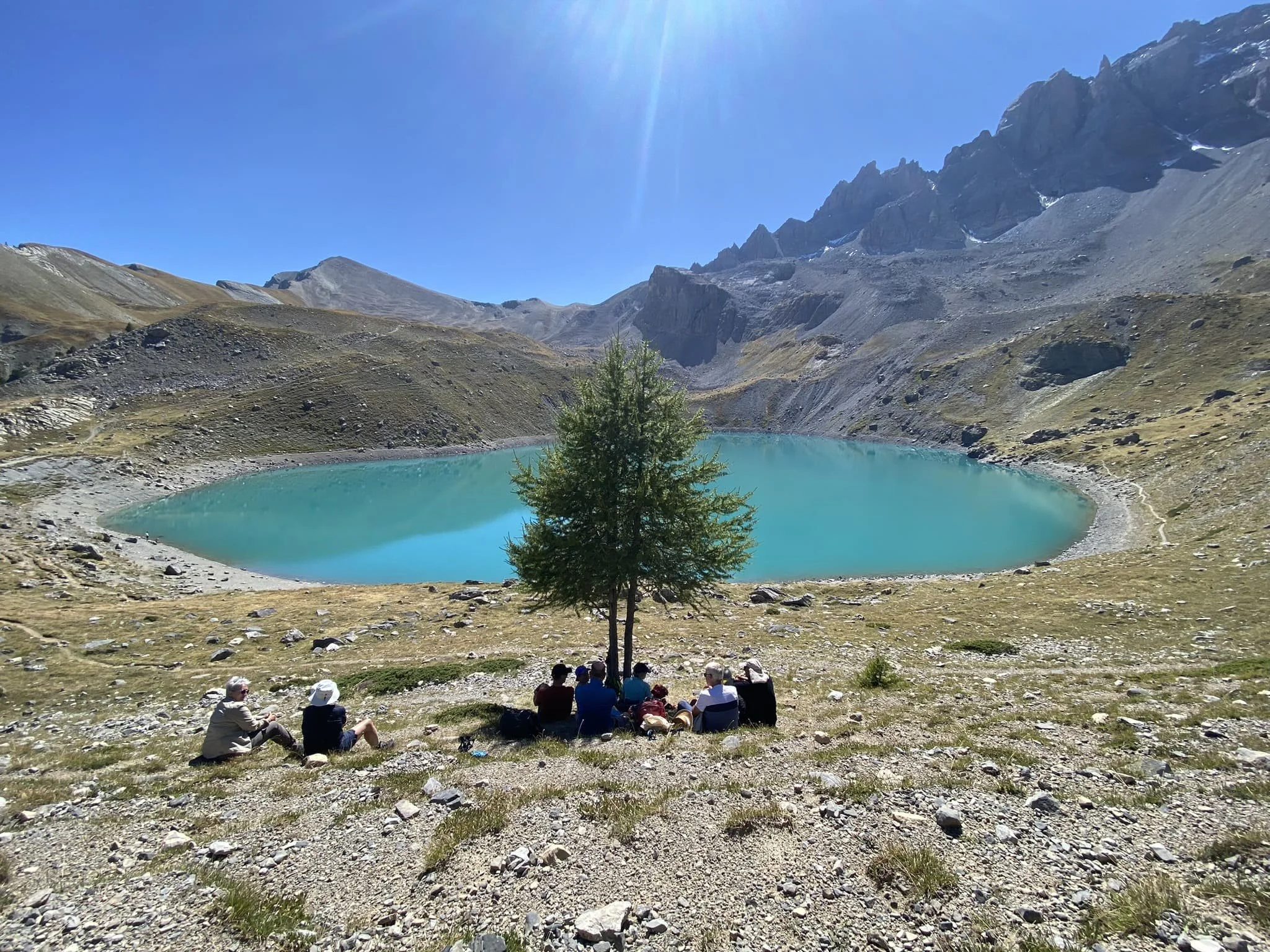 Groupe de personnes assises sous un arbre près d'un lac de montagne turquoise, entouré de reliefs rocheux et de montagnes escarpées en arrière-plan, sous un ciel bleu ensoleillé.