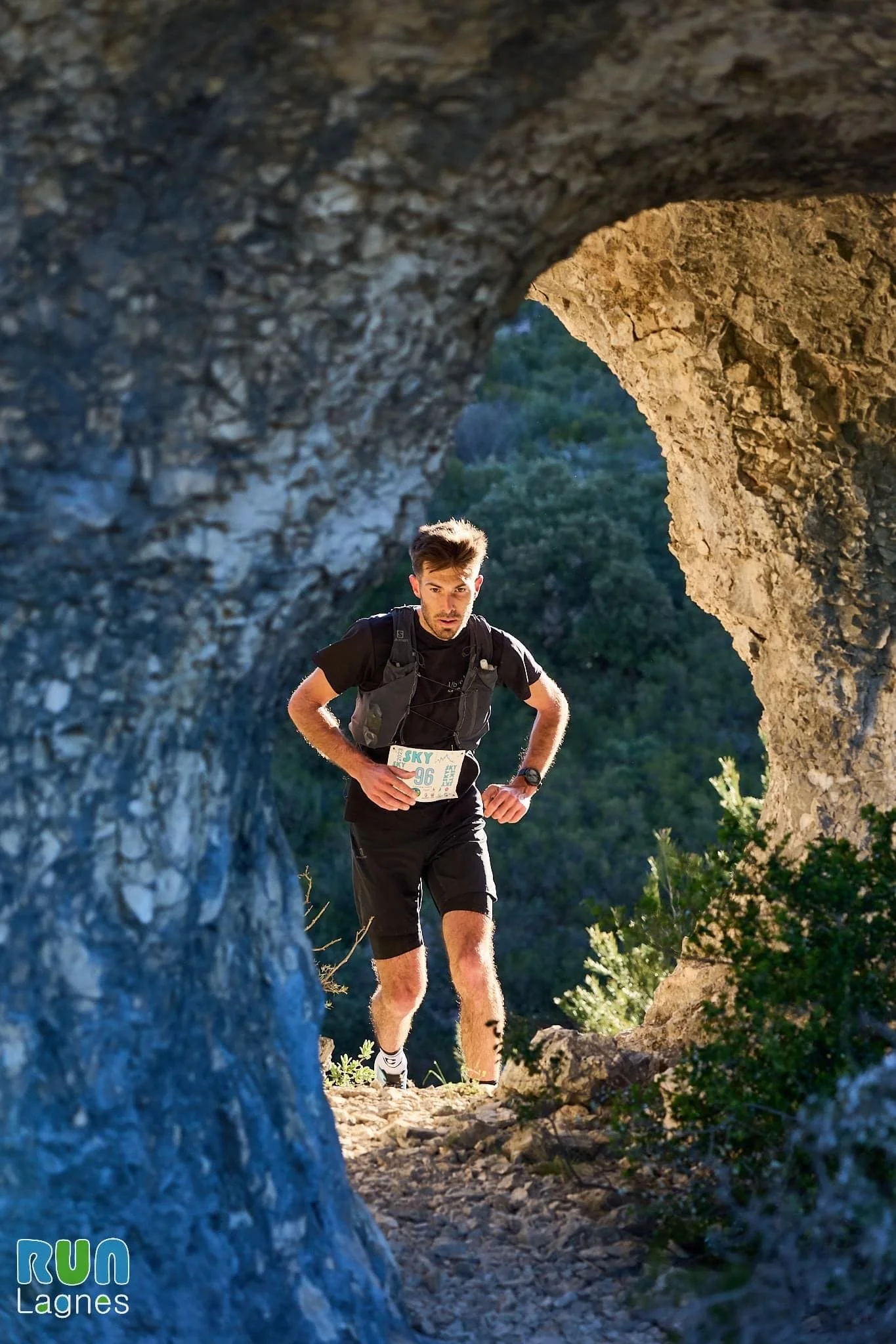 Un homme court sur un chemin escarpé vu à travers un trou dans une roche, avec un arrière-plan de végétation verte et un ciel clair.