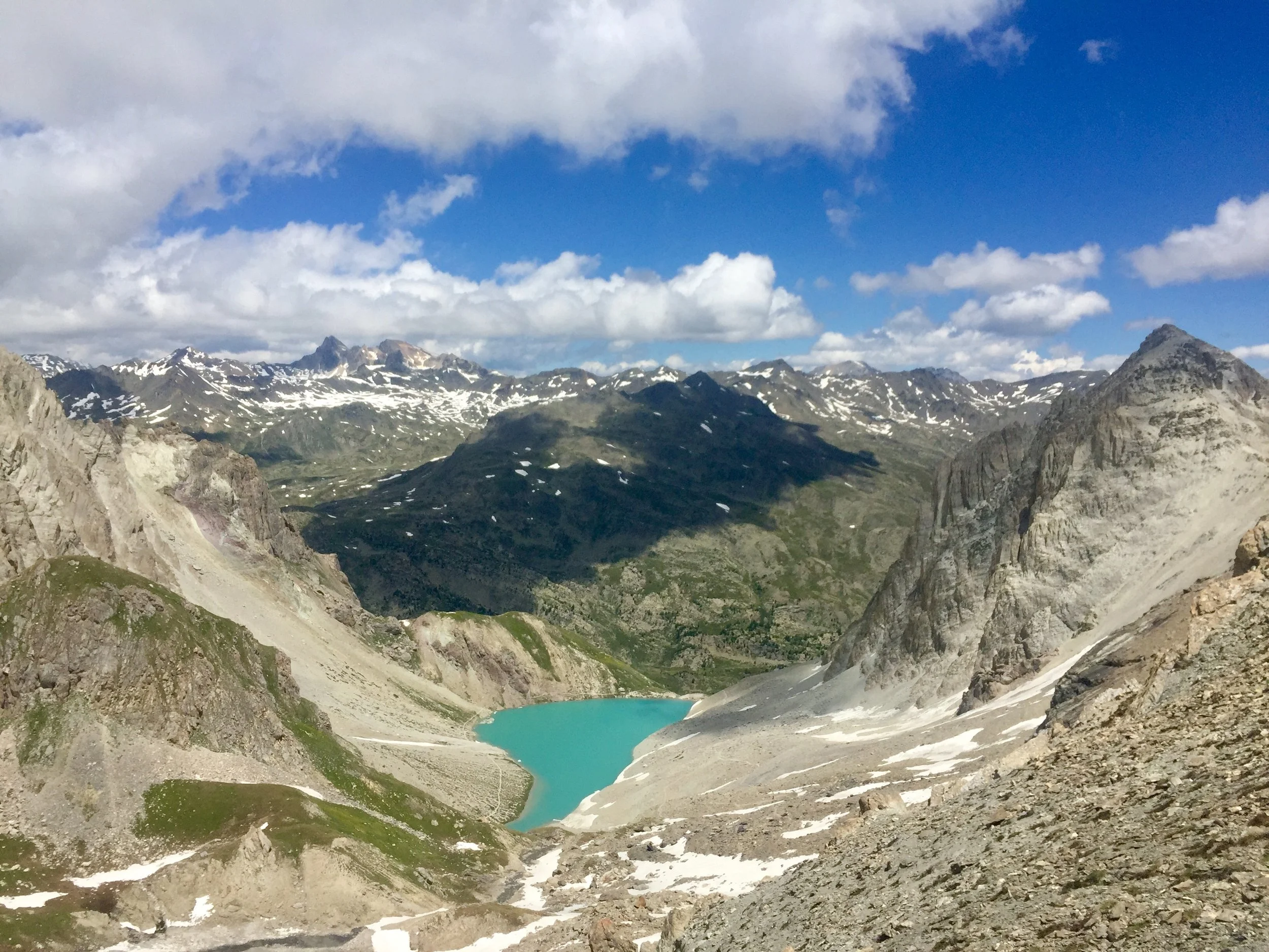 Paysage de montagnes avec lac turquoise au centre, sommets enneigés et ciel bleu avec nuages.