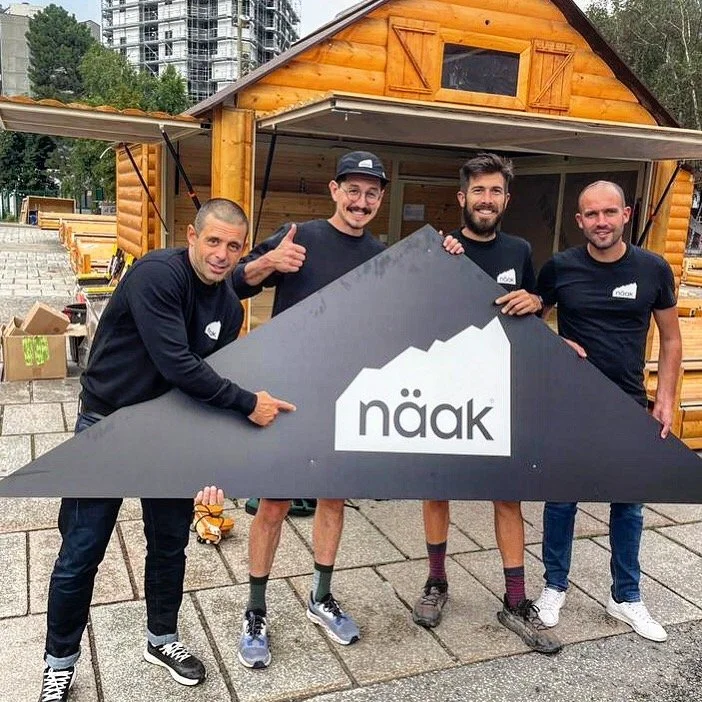 Quatre hommes souriants tenant une grande enseigne noire avec le logo 'näak' devant une petite maison en bois, dans un environnement urbain avec des bâtiments en construction ou en cours de rénovation.