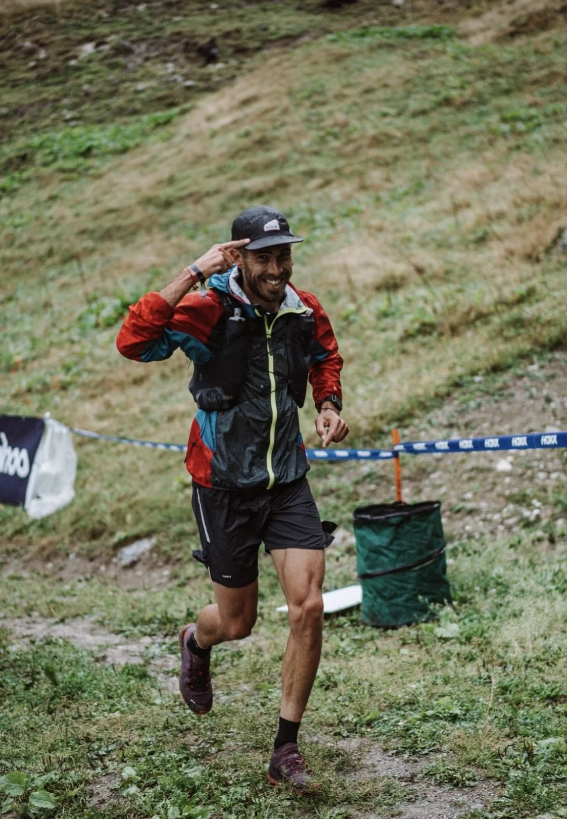 Un homme souriant en course à pied dans un sentier de montagne, portant un chapeau, un manteau de sport, et un sac à dos.