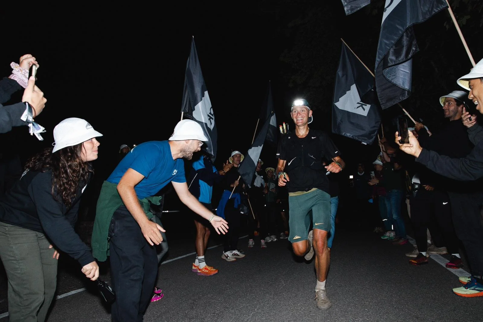 Un homme courant la nuit lors d'une course d'ultra-marathon, entouré de spectateurs applaudissant et prenant des photos.