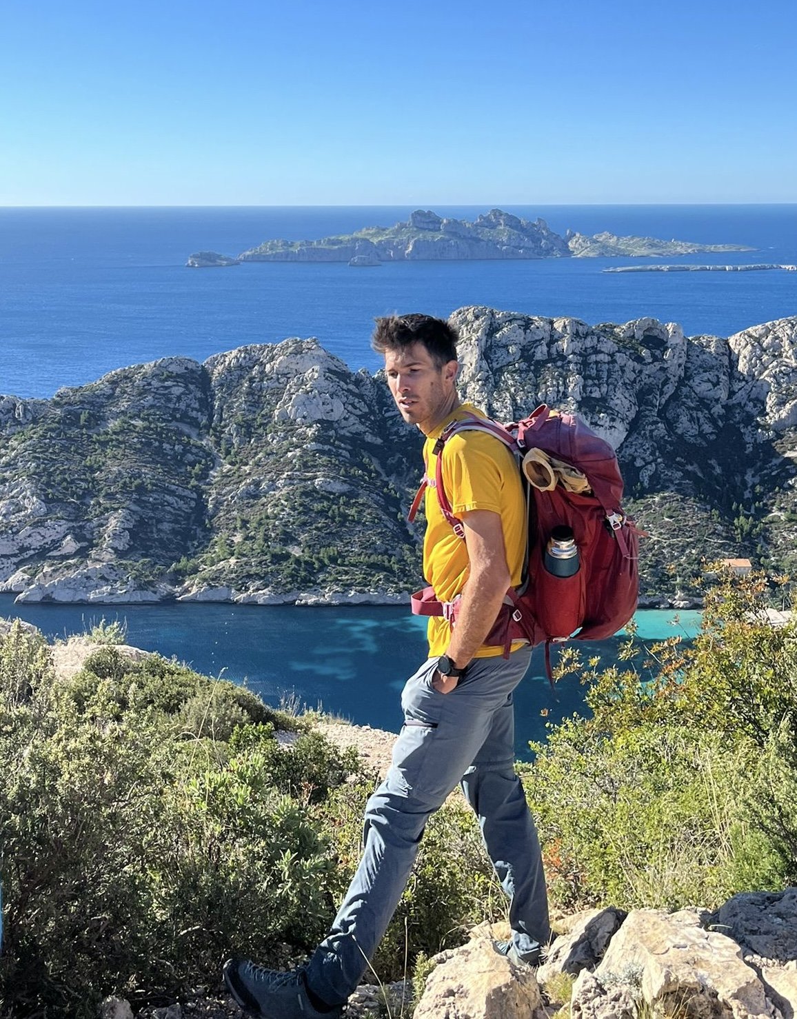 Homme en randonnée avec un sac à dos jaune, au bord de la mer avec des rochers et des îles en arrière-plan, ciel bleu clair.