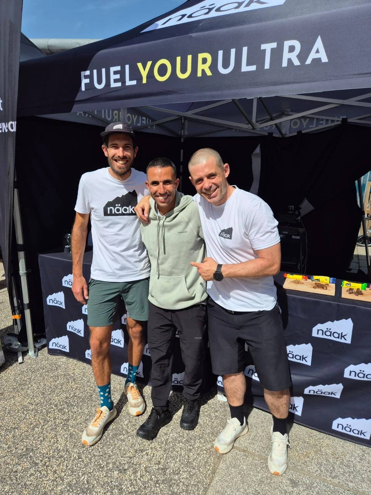 Trois hommes souriants posant sous une tente noire avec le texte "FUEL YOUR ULTRA". Deux portent des T-shirts blancs avec le logo Näak, le troisième porte un sweat vert. Au fond, une table avec des barres énergétiques.