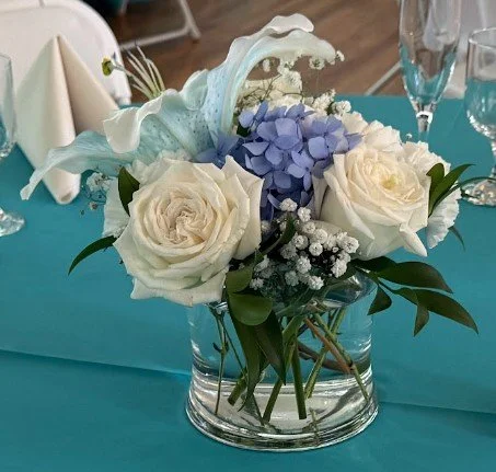 Floral centerpiece with white roses, blue hydrangeas, baby's breath, and greenery on a teal tablecloth at a formal event.