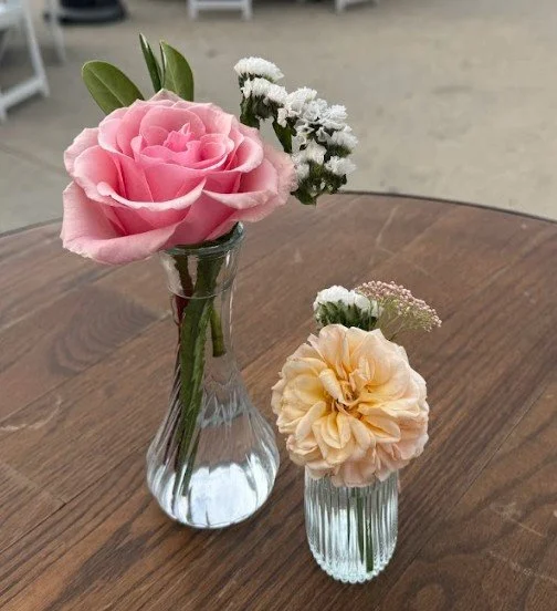 Two small glass vases with flowers on a wooden table. The larger vase has a pink rose, white baby's breath, and green leaves. The smaller vase has a peach-colored carnation and some small white flowers.