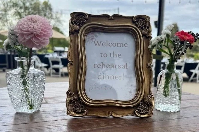 Freestanding ornate gold picture frame on a table with a sign inside that says 'Welcome to the rehearsal dinner!' Flanked by two glass vases with pink and white flowers. Outdoor setting with chairs and trees in the background.
