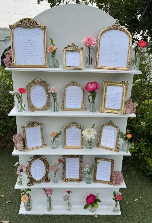A white multi-shelf display with framed quotes and small flower vases containing pink, white, and peach flowers, set outdoors against green foliage.