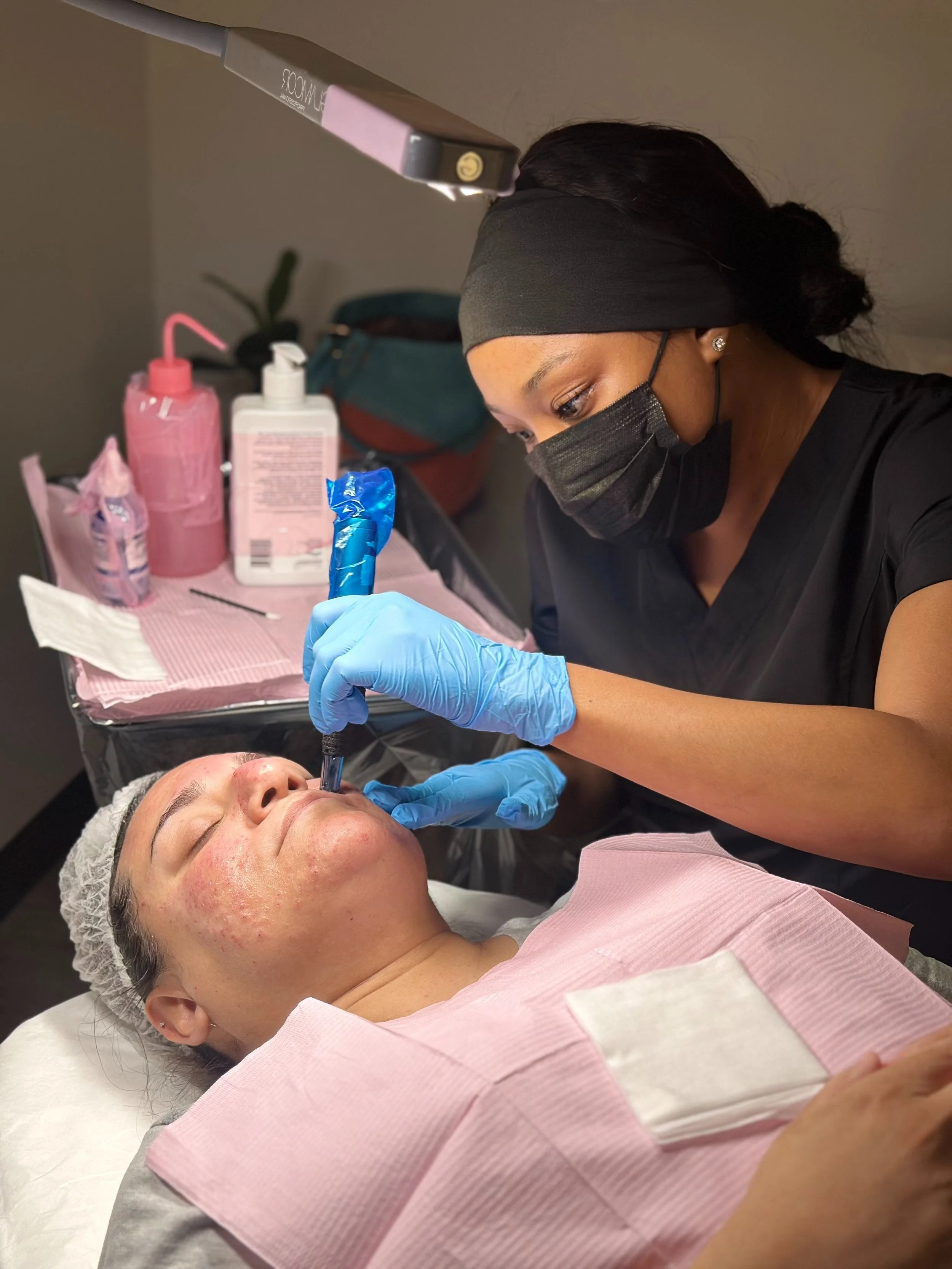 Skincare specialist performing microneedling on a woman lying down, wearing a hair cap and pink protective sheet, with skincare products nearby.