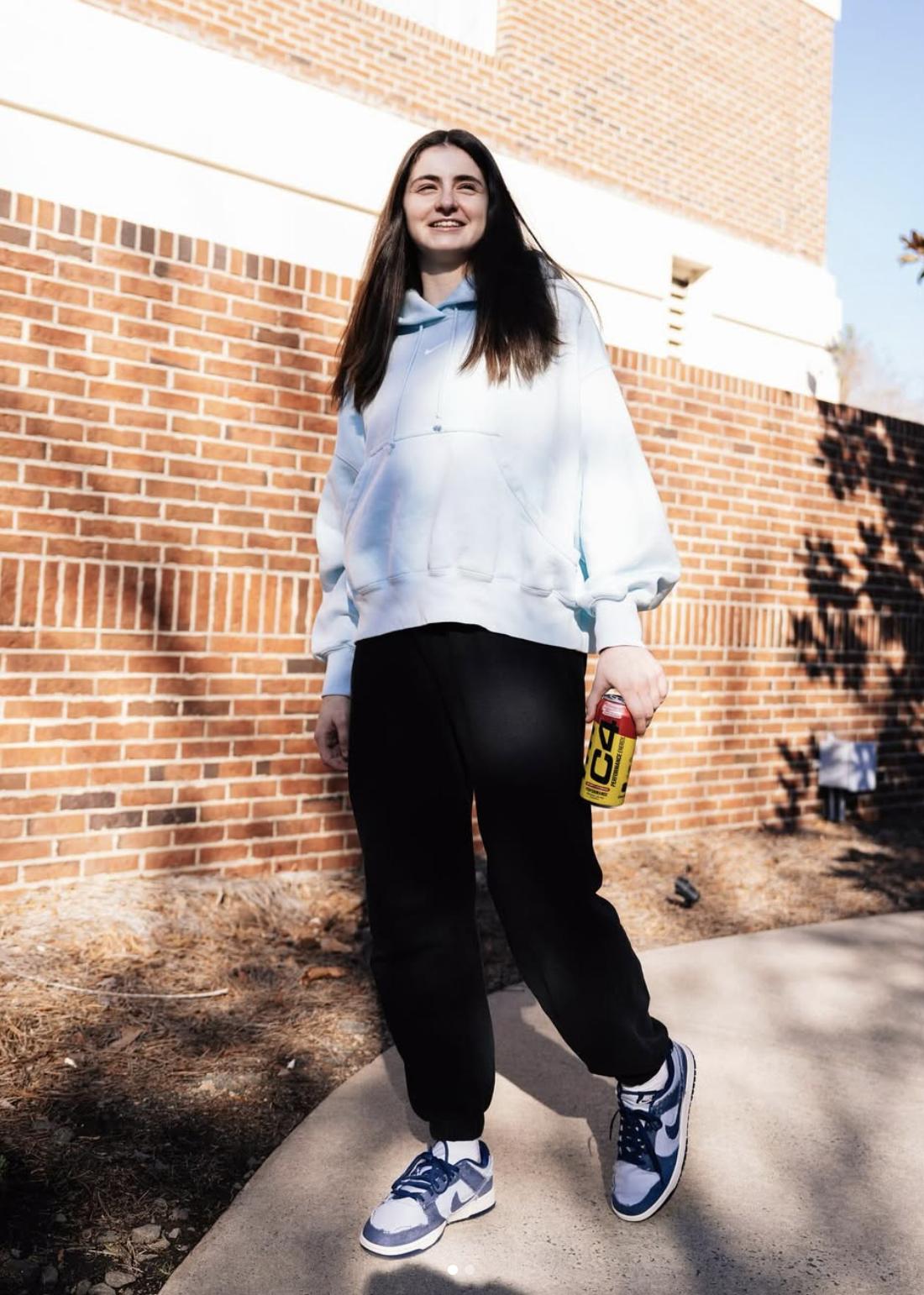 Young woman with long dark hair smiling, wearing a white hoodie, black sweatpants, and blue and white sneakers, holding a red and yellow canned beverage, standing outside against a brick wall on a sunny day.