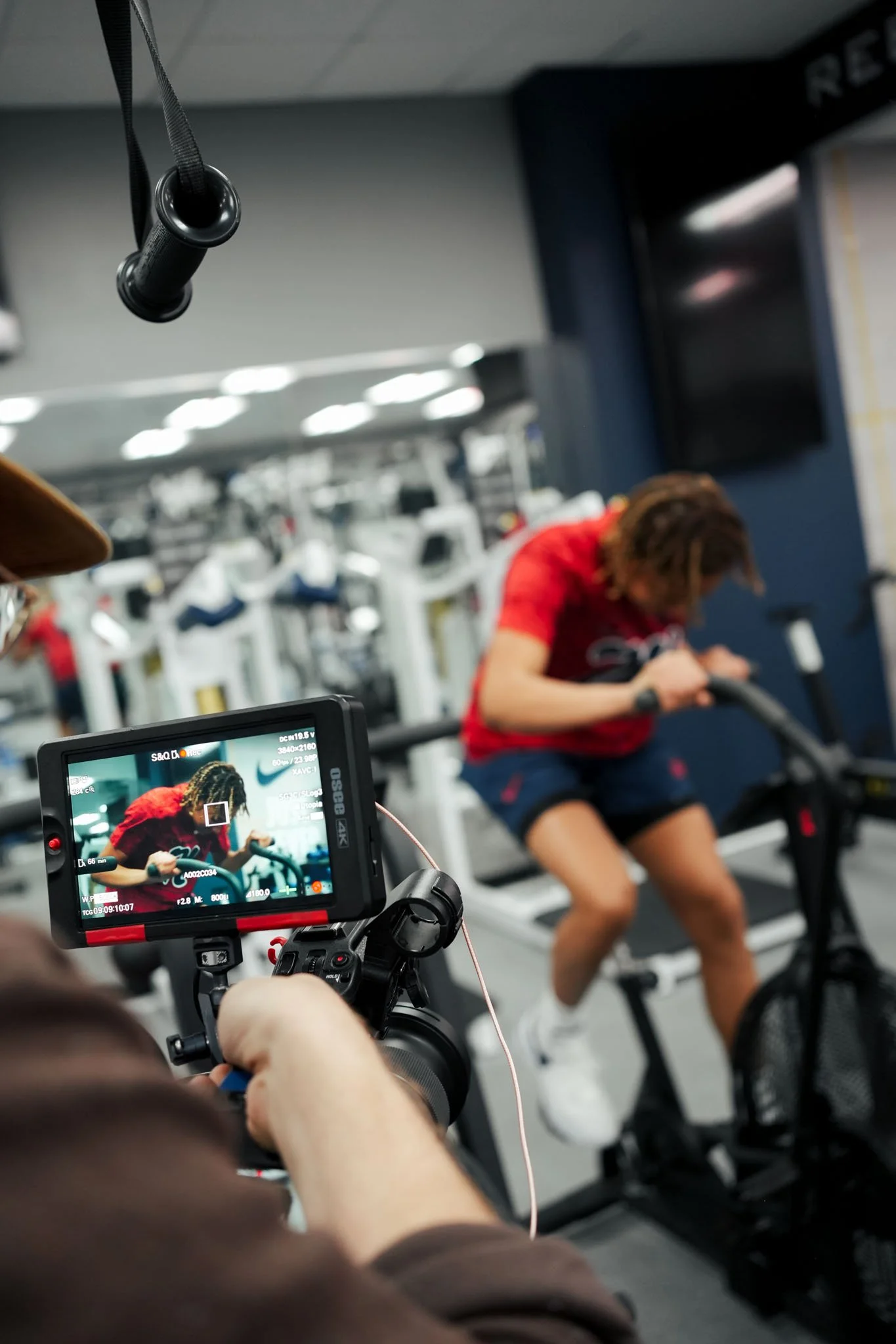 A person filming a woman on a rowing machine at a gym with a camera and monitor capturing the scene.