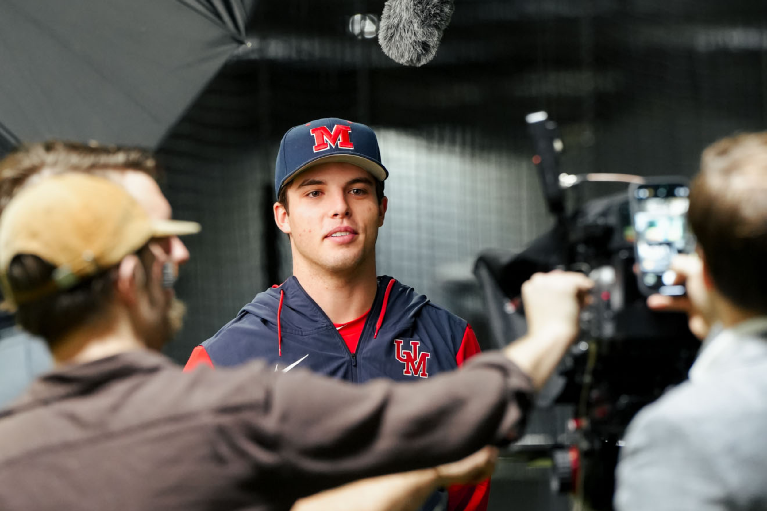 A young man in a baseball cap and sports jacket being filmed or interviewed by crew members in an indoor setting.