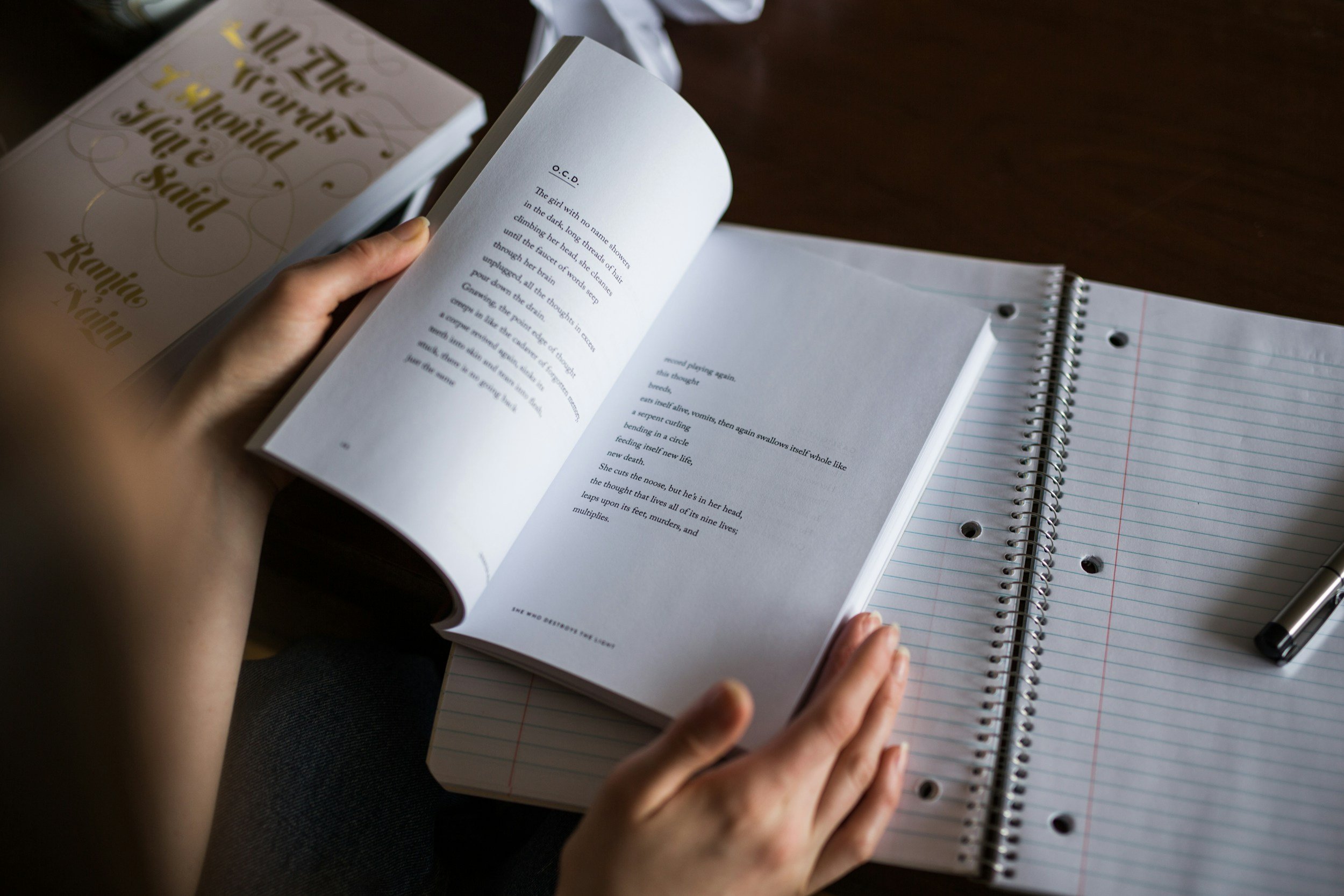 Person flipping through a small book or booklet, with lined spiral notebook and pen on a wooden surface.