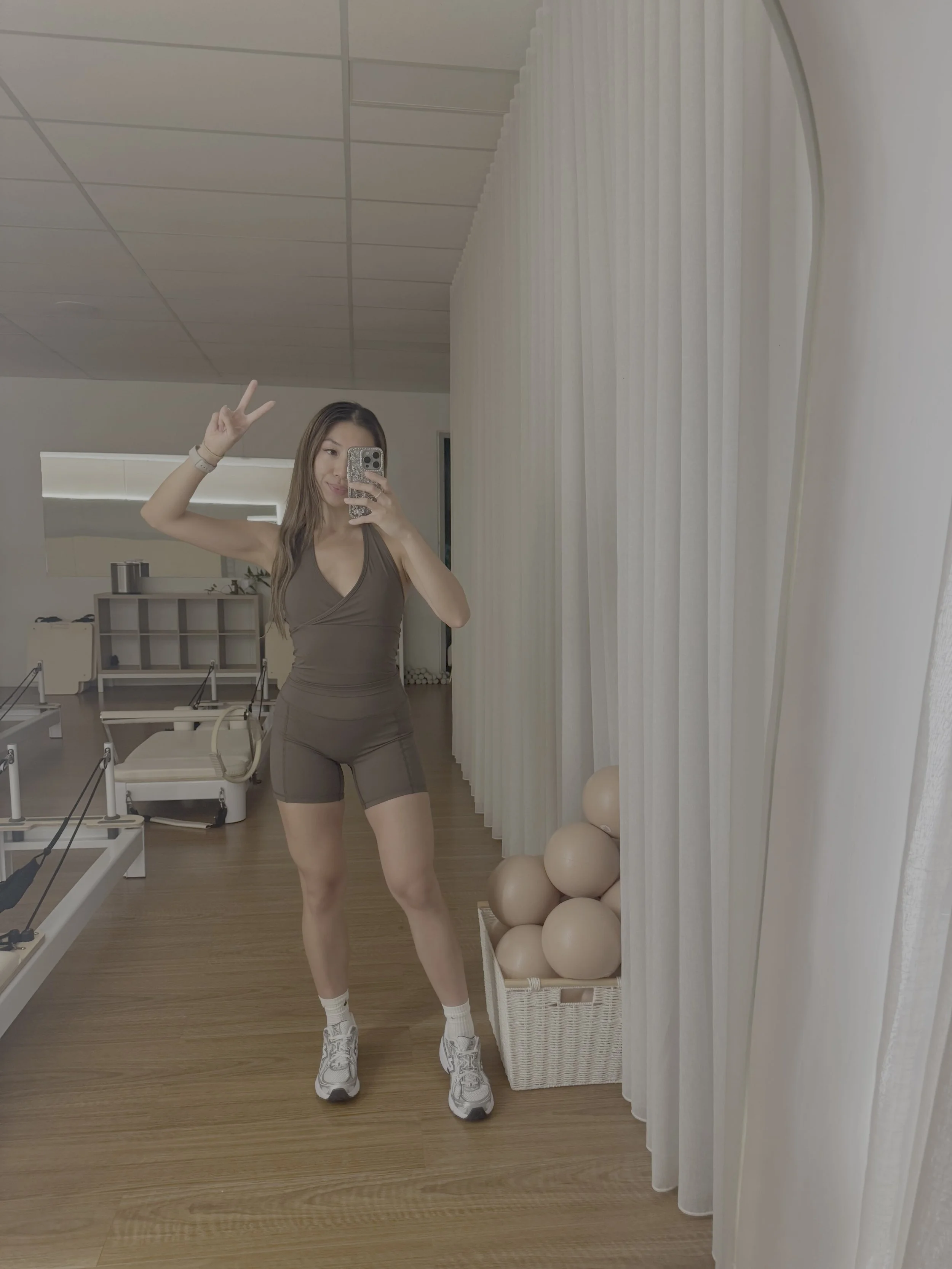 A woman taking a mirror selfie with a peace sign, wearing a brown workout outfit and sneakers in a fitness studio with medicine balls and yoga mats.