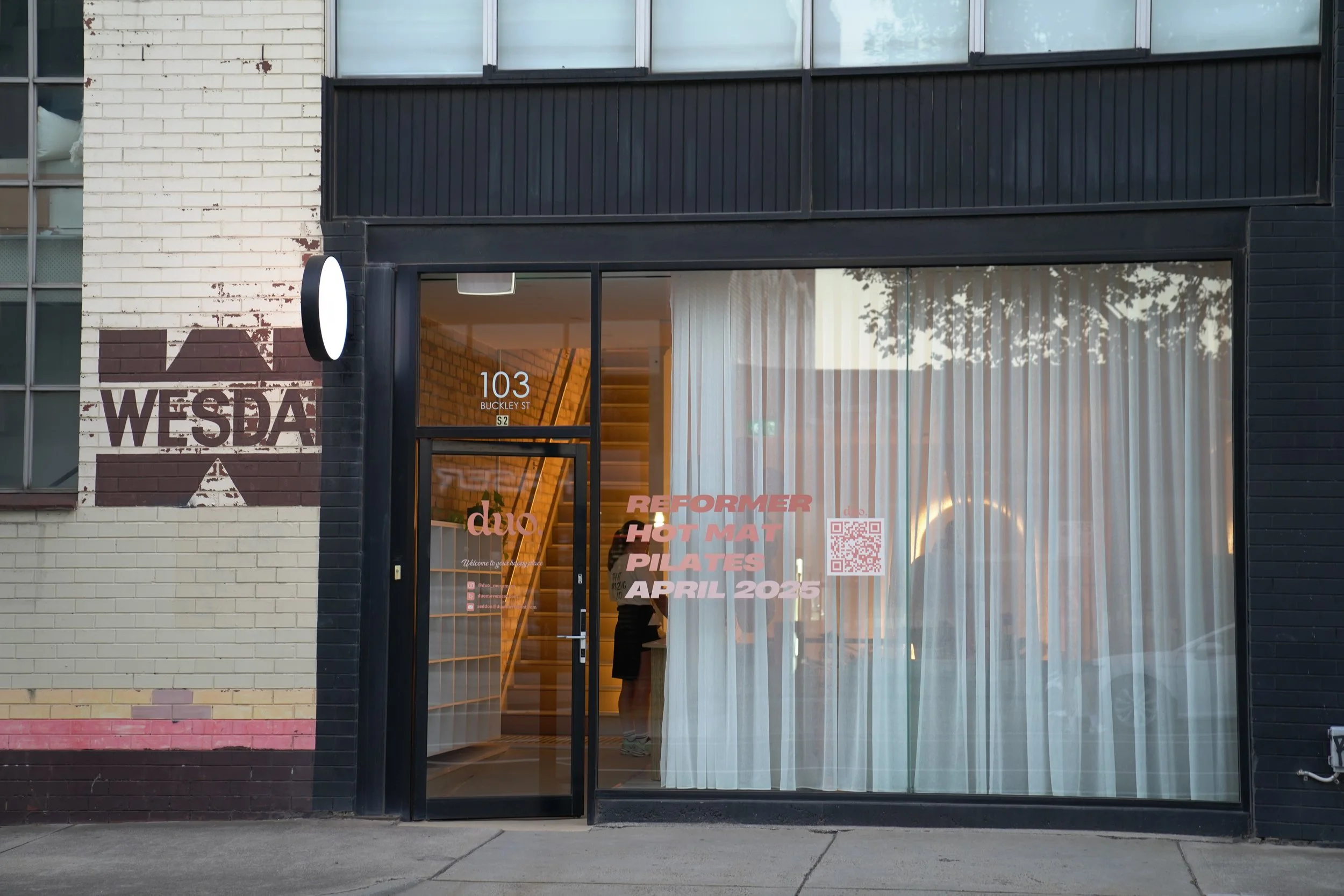 storefront with the address 103 BUCKLEY ST, black frame glass door, white brick wall with WESDA painted logo, inside a staircase, people, sheer curtains, and advertising text for reformer hot mat Pilates in April 2025 with a QR code.