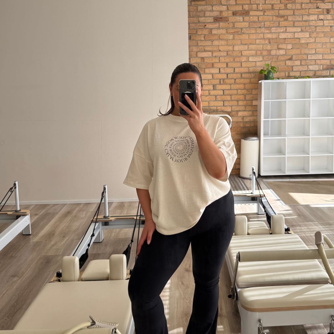 A woman taking a mirror selfie in a fitness or Pilates studio. She is wearing a beige oversized t-shirt and black leggings, with exercise equipment and a brick wall in the background.