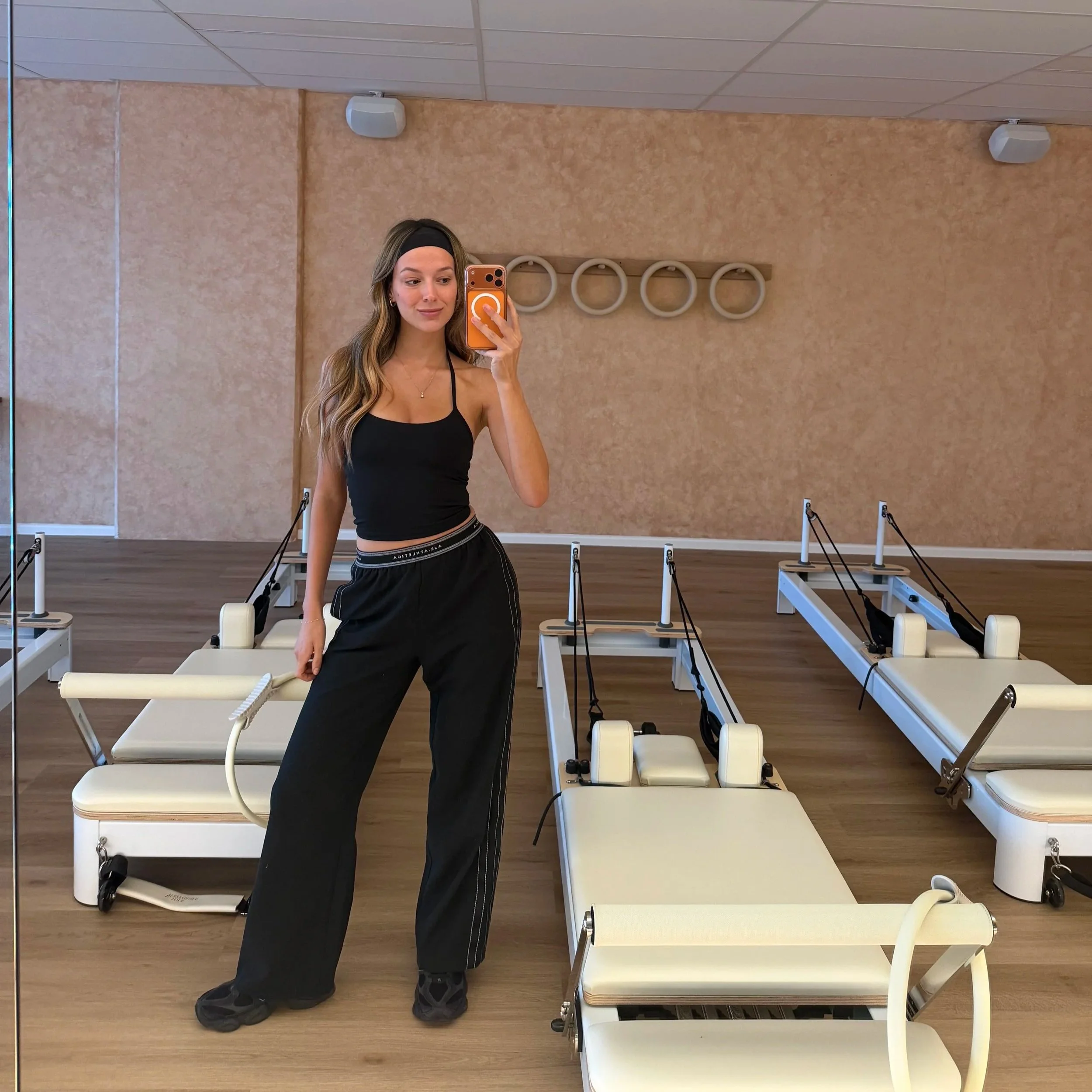 A woman taking a mirror selfie in a Pilates studio with Pilates reformer machines in the background.