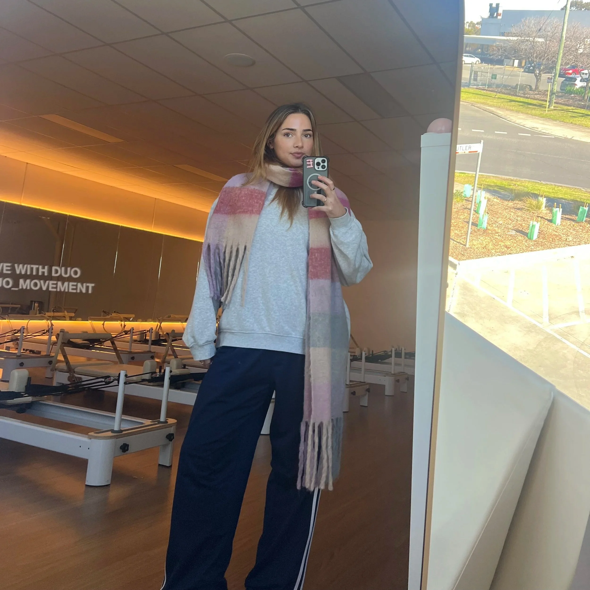 A woman taking a selfie in a mirror, wearing a light gray sweater, navy pants, and a pink, beige, and white plaid scarf inside a modern building. Outside the window, there's a parking lot, trees, and a clear blue sky.