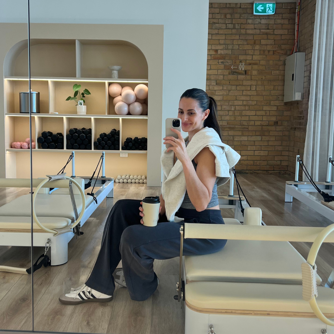 A woman sitting in a gym or fitness studio taking a selfie in the mirror while holding a coffee cup, with gym equipment and shelves with exercise balls and dumbbells in the background.