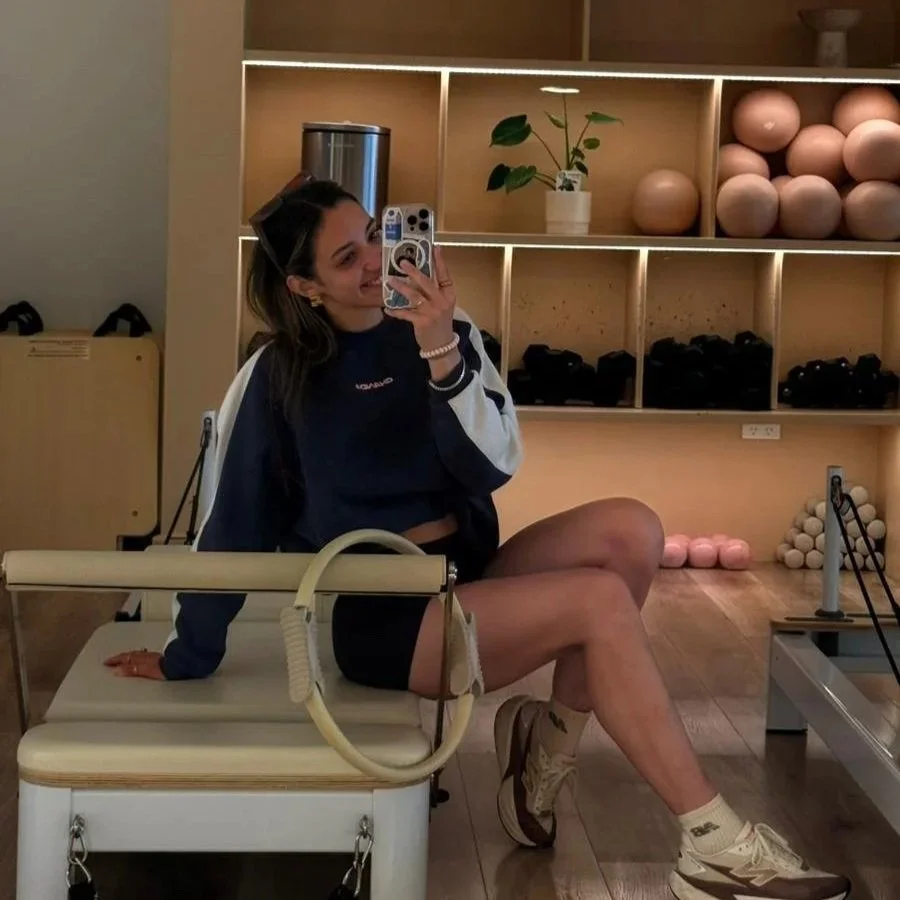 Young woman taking a selfie in a fitness or physical therapy room, sitting on a treatment table, with gym equipment and decorative objects in the background.