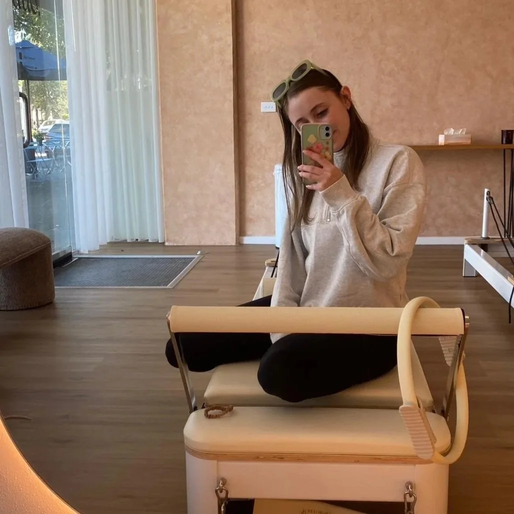 A young woman taking a selfie in a mirror inside a room with beige walls and wooden flooring. She has sunglasses on her head, is wearing a beige sweater and black pants, and is seated on a white rolling cart. There is a large window with white curtains, and outside, a patio with an umbrella and parked cars are visible.