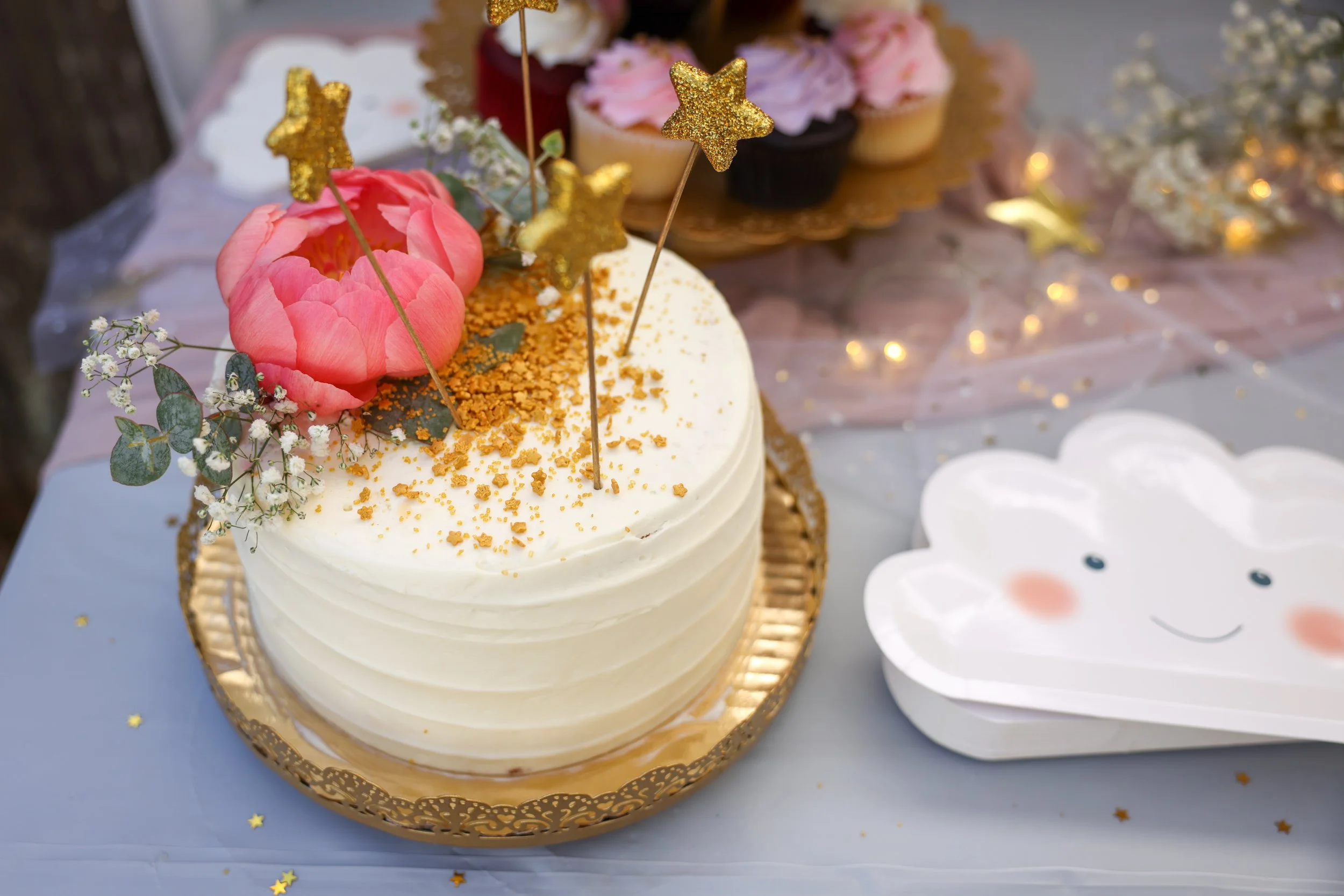 A decorated white cake with pink flower, gold star toppers, and gold sprinkles, on a gold tray, with cupcakes in the background.