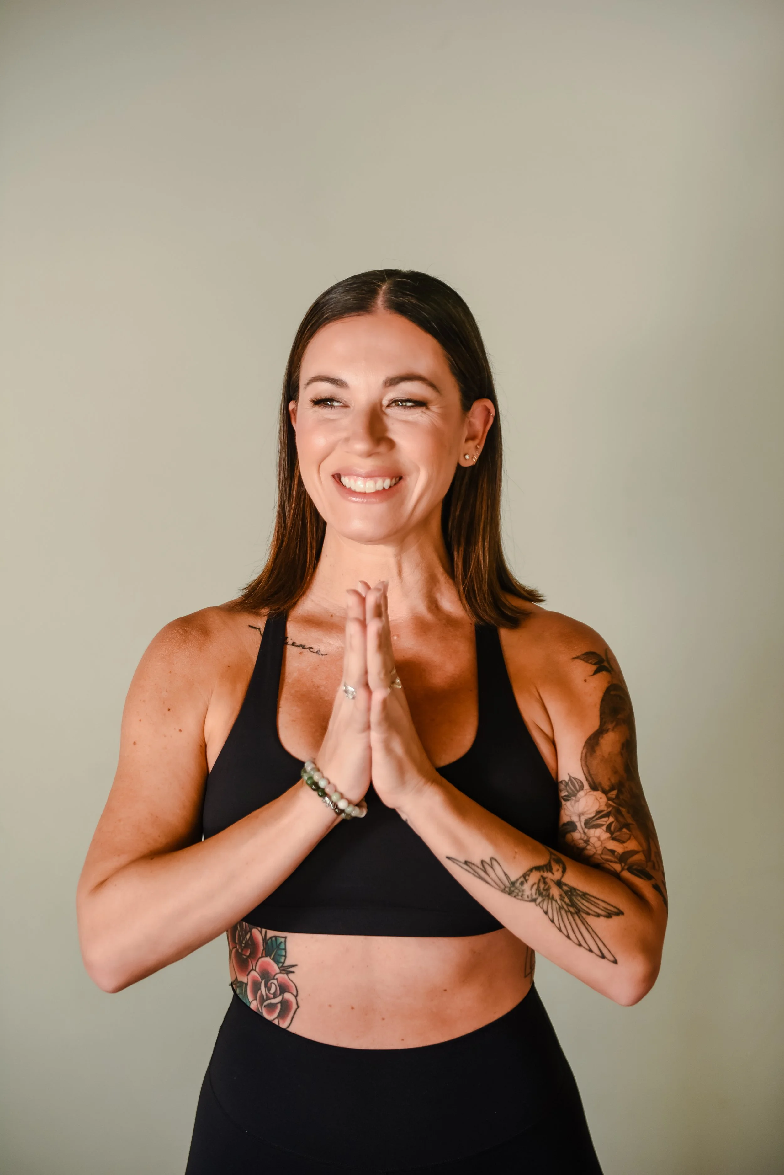 A woman with long, wavy brown hair smiling, wearing a black tank top, standing in front of a stone wall.