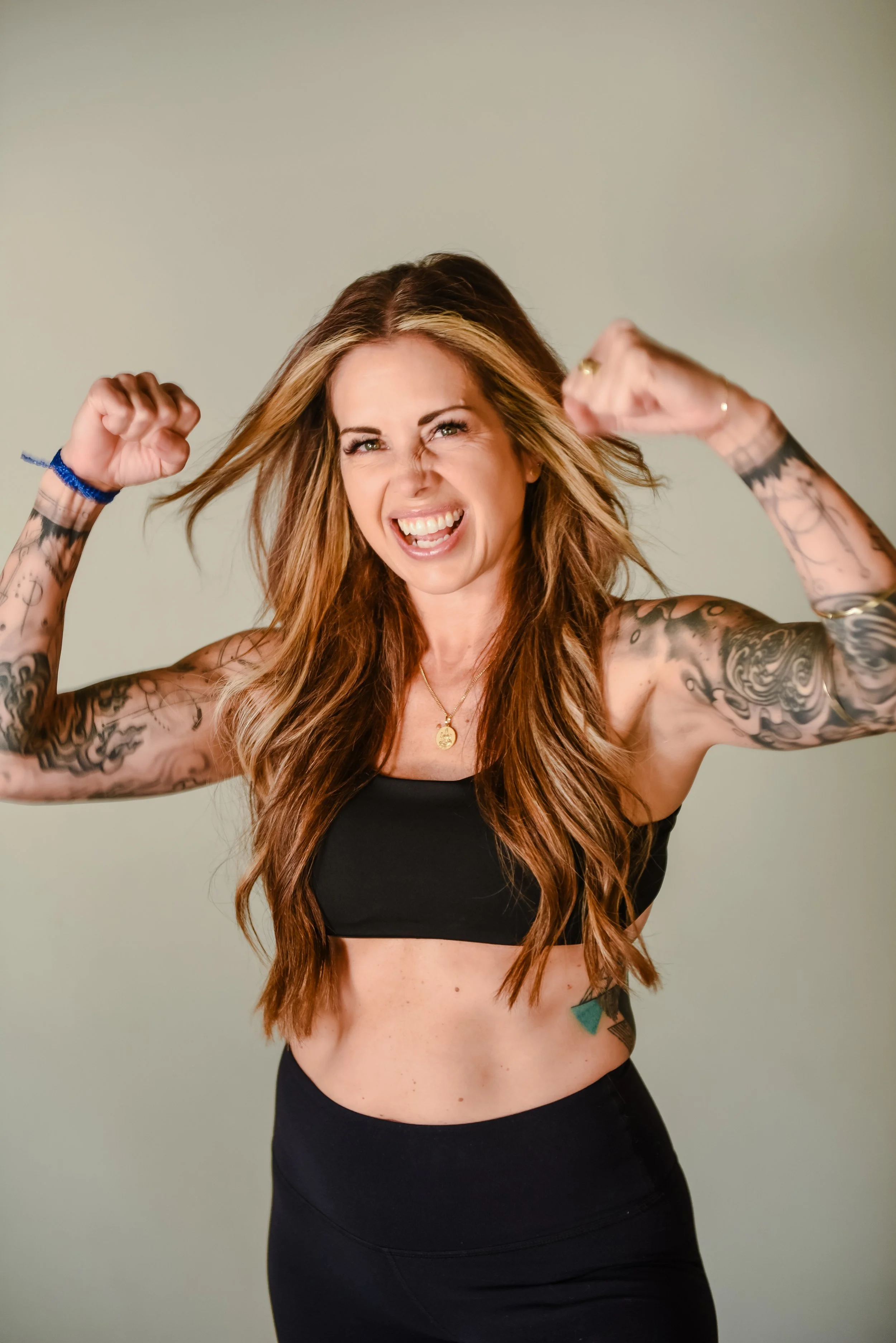 A smiling woman with tattoos wearing a black sports bra and black high-waisted pants, standing against a brick wall.