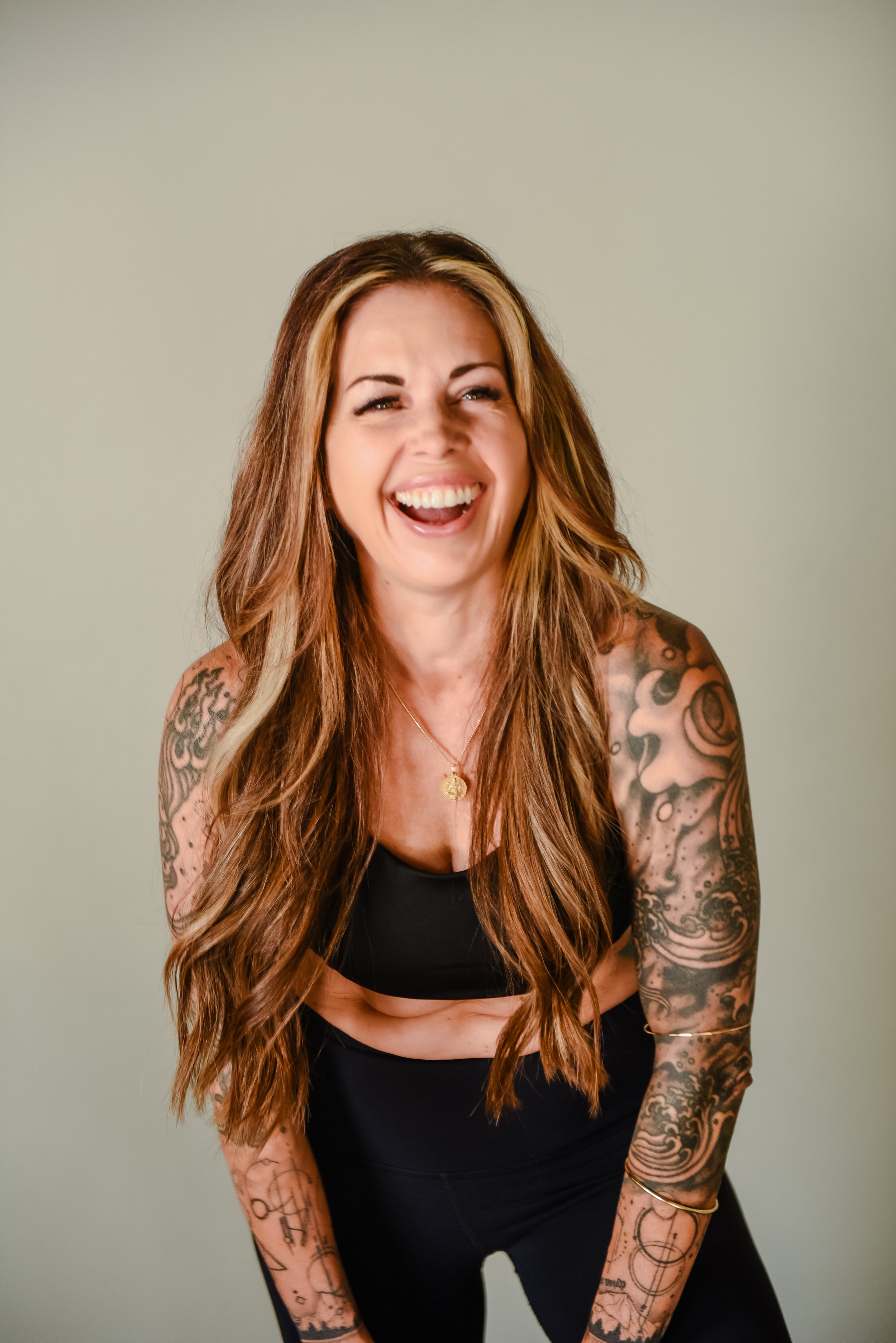 A smiling woman with tattoos wearing a black sports bra and black high-waisted pants, standing against a brick wall.