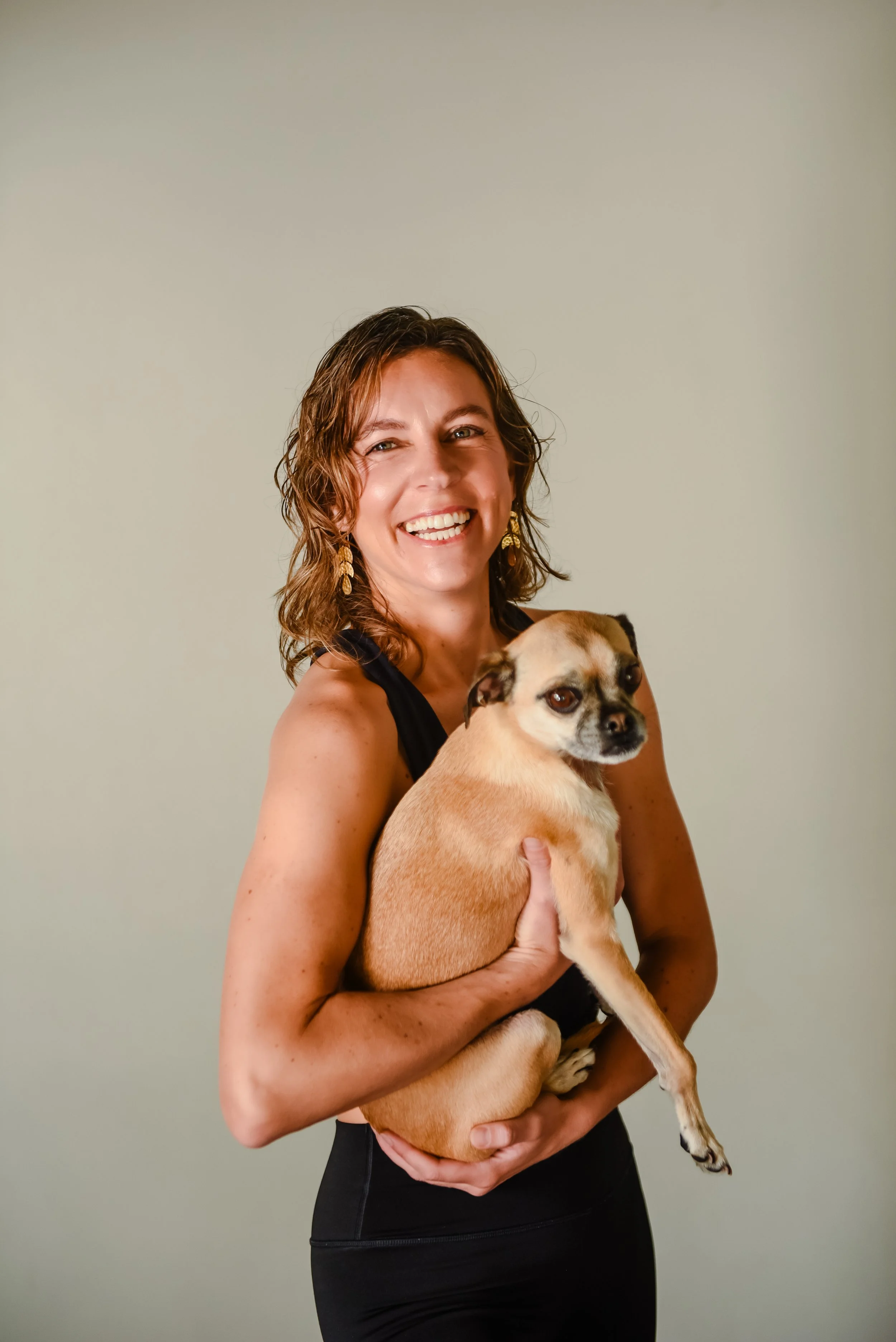 A woman with wet, dark hair smiling in front of a stone wall, wearing a black tank top and earrings with blue stones.