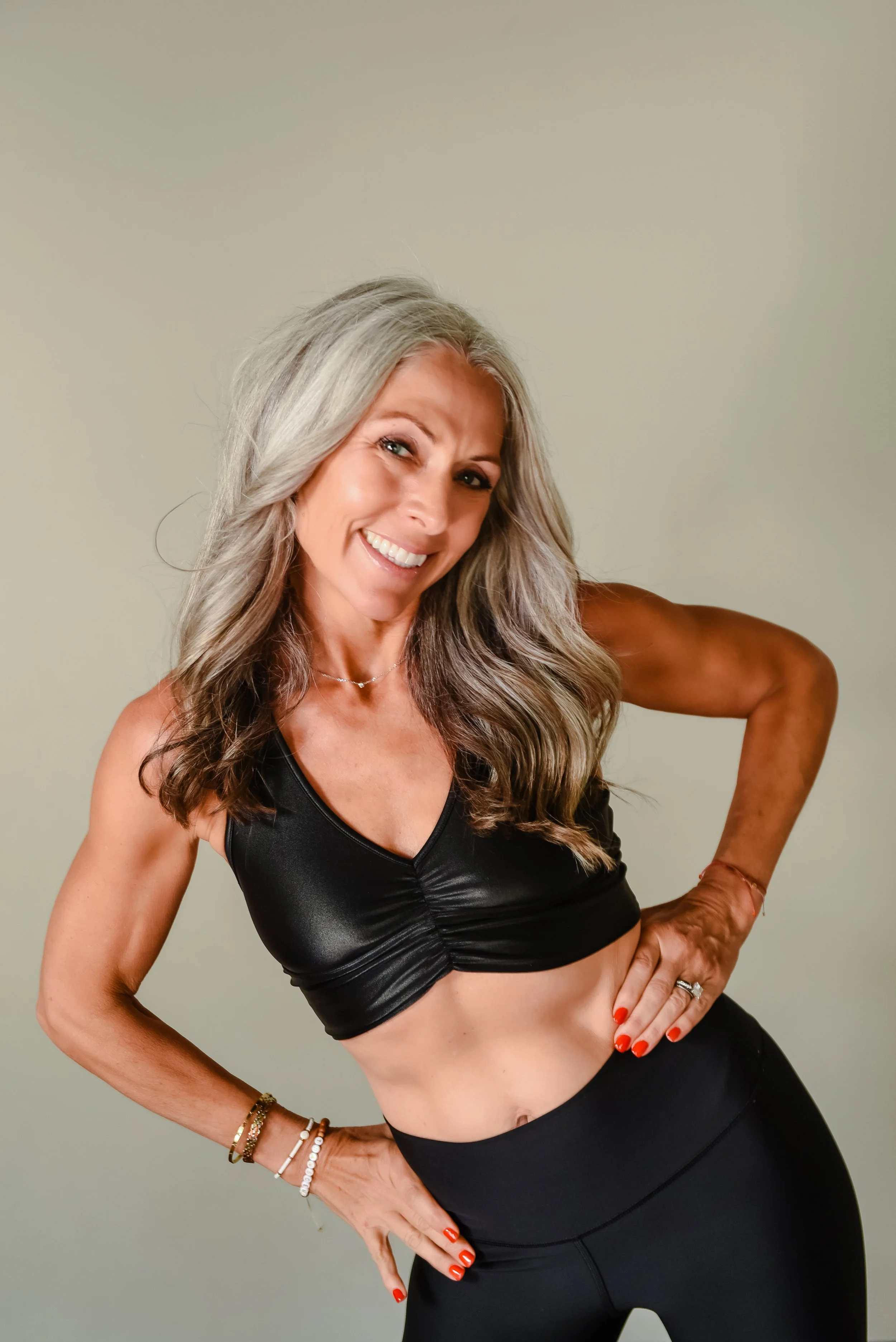 A woman with gray hair styled in loose waves, wearing a white tank top, holding her hands together in a prayer gesture, with a calm expression, standing against a dark background.