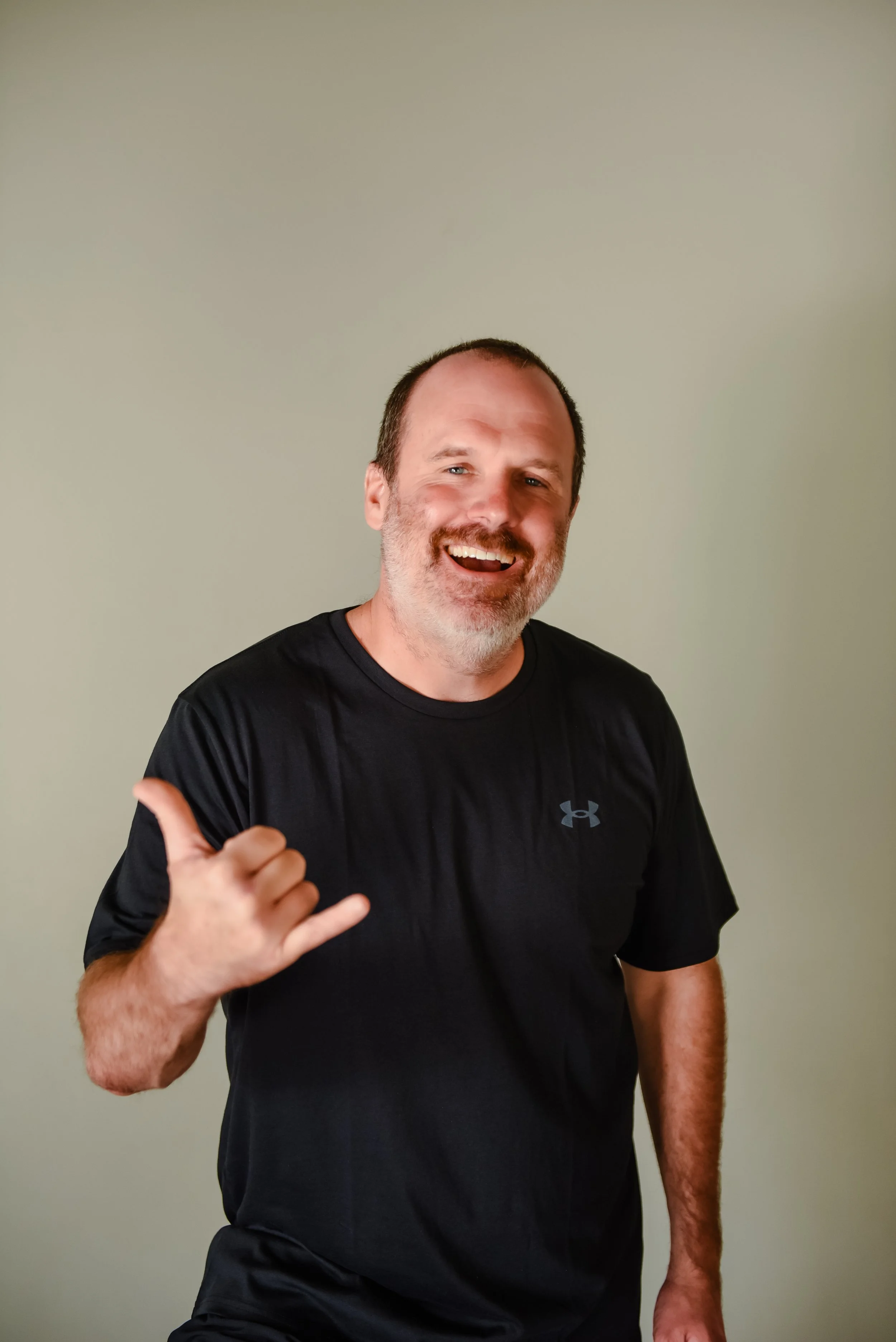 Smiling man with a beard and short dark hair standing in front of a stone wall, wearing a black T-shirt.
