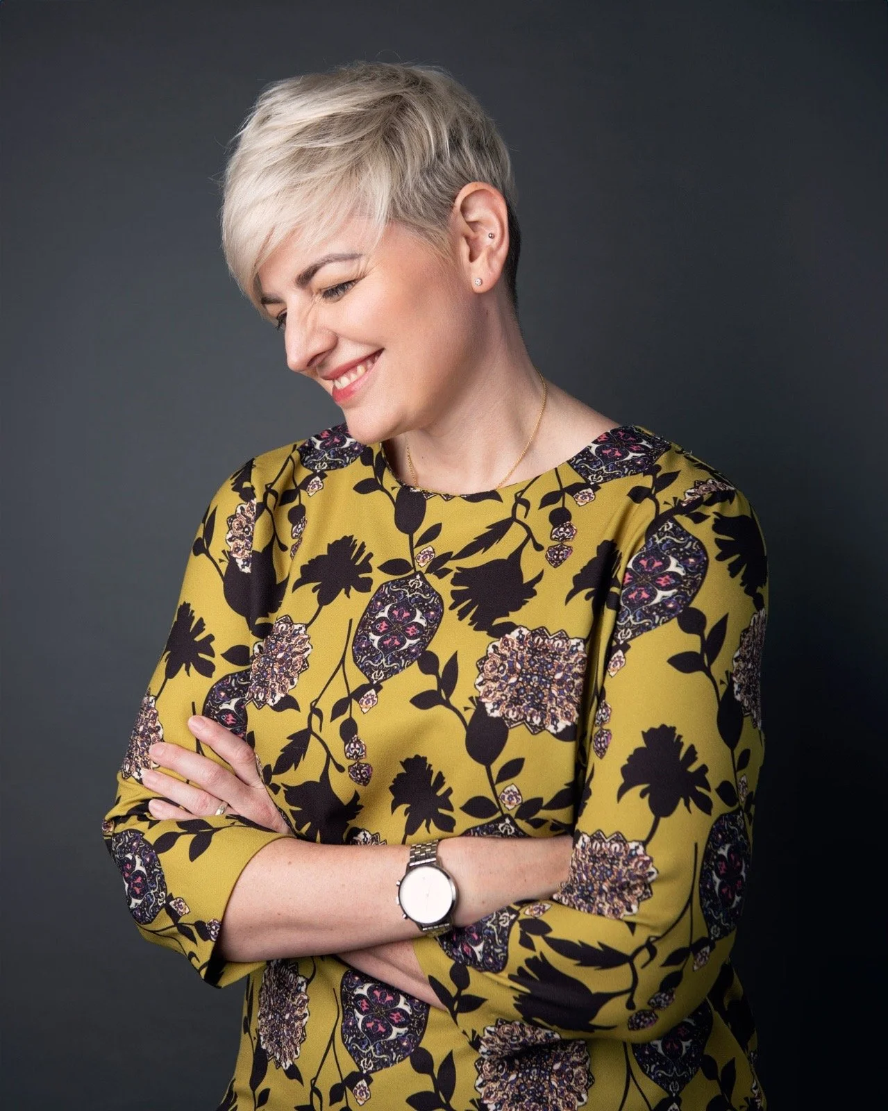 A woman with short blonde hair in a pixie cut, wearing a yellow dress with black and purple floral patterns. She has earrings, a necklace, a watch, and is smiling with eyes closed, arms crossed, against a dark background.