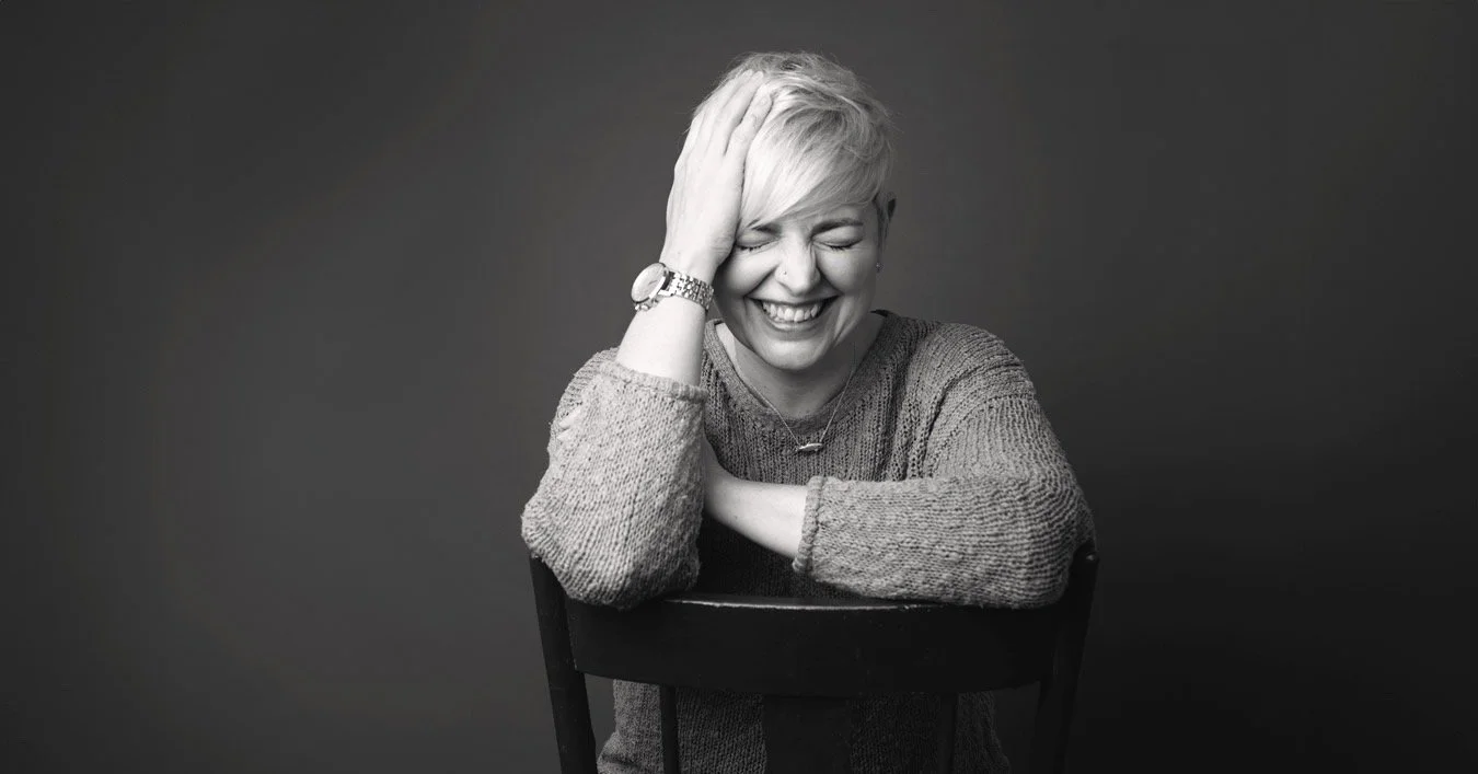 A woman with short, light-colored hair laughing with her eyes closed, wearing a sweater, watch, and necklace, sitting at a table against a dark background.