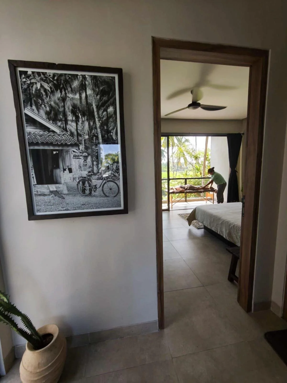 View of a bedroom with a bed, a woman giving a massage, and large glass doors opening to a balcony with trees outside. A black and white framed photo hang on the wall.