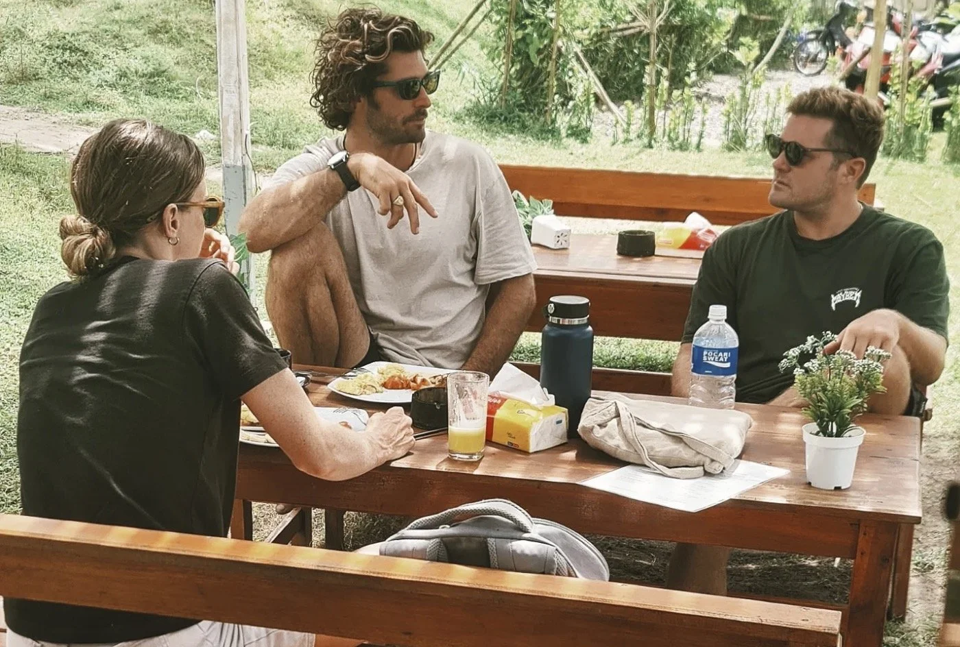Three people sitting at a wooden picnic table outdoors, engaged in conversation, with food and drinks on the table.