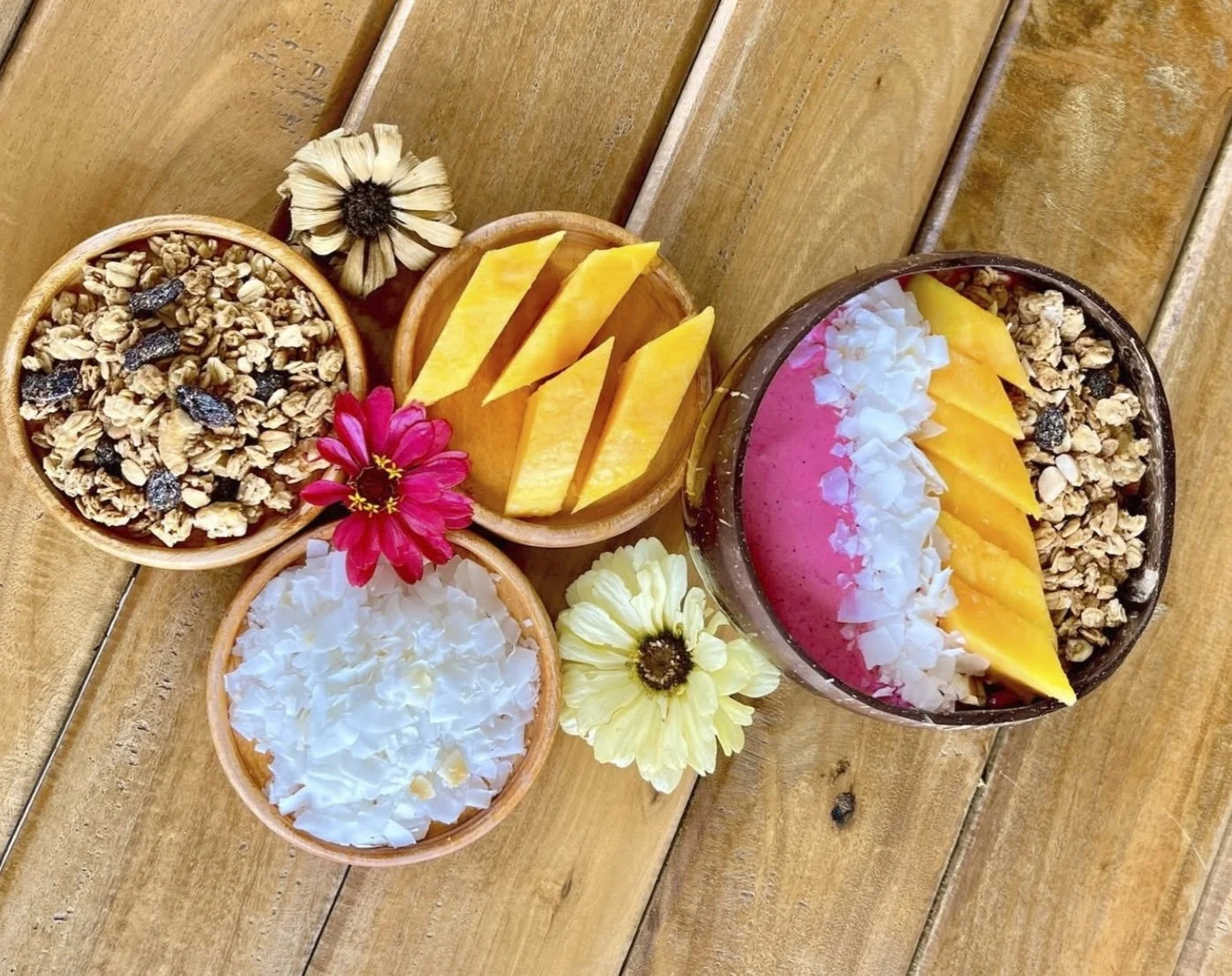 A smoothie bowl topped with shredded coconut, pineapple slices, granola with dried fruits, and fresh flowers on a wooden table.