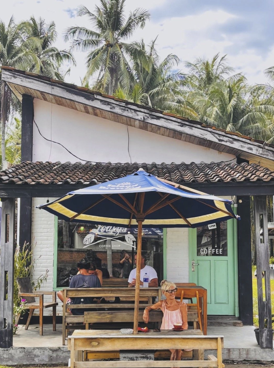 People enjoying coffee outside of a small coffee shop with a turquoise door and green window trim. The shop has a wooden porch with a bench and a large blue umbrella. There are palm trees in the background.