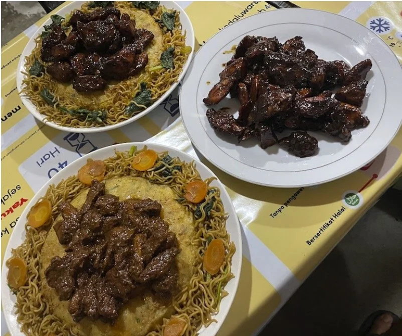 Three plates of Indonesian food on a table: one plate of deep-brown beef rendang with noodles, another of braised beef ribs, and a third of beef rendang with rice and vegetables.